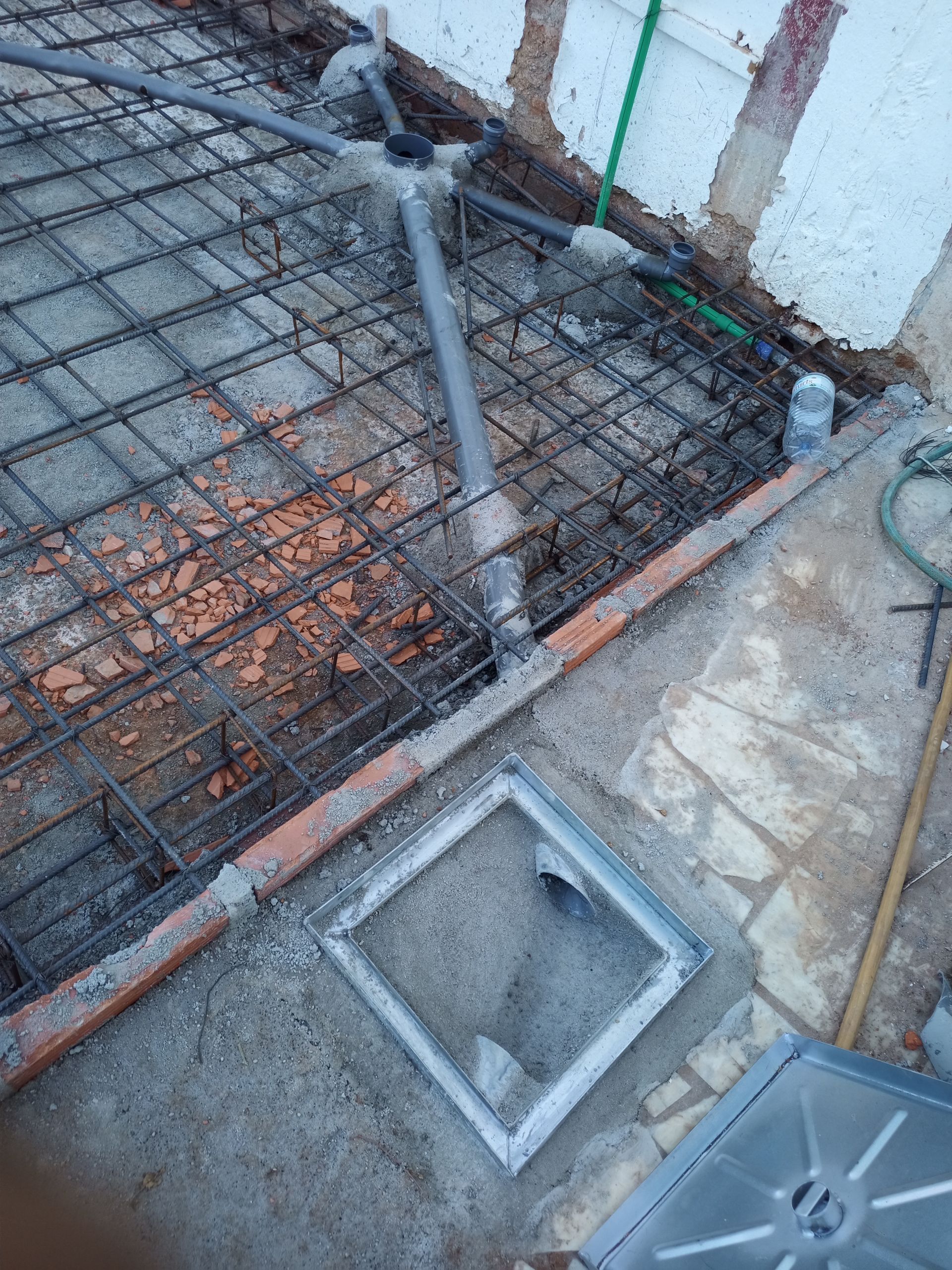 Canteiro de obras com malha de vergalhões, tubos de PVC e dreno de concreto com acesso aberto.