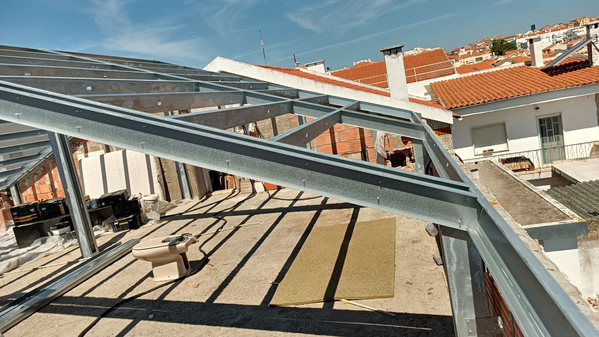 Estrutura metálica de um telhado em construção, com edifícios circundantes e céu azul.