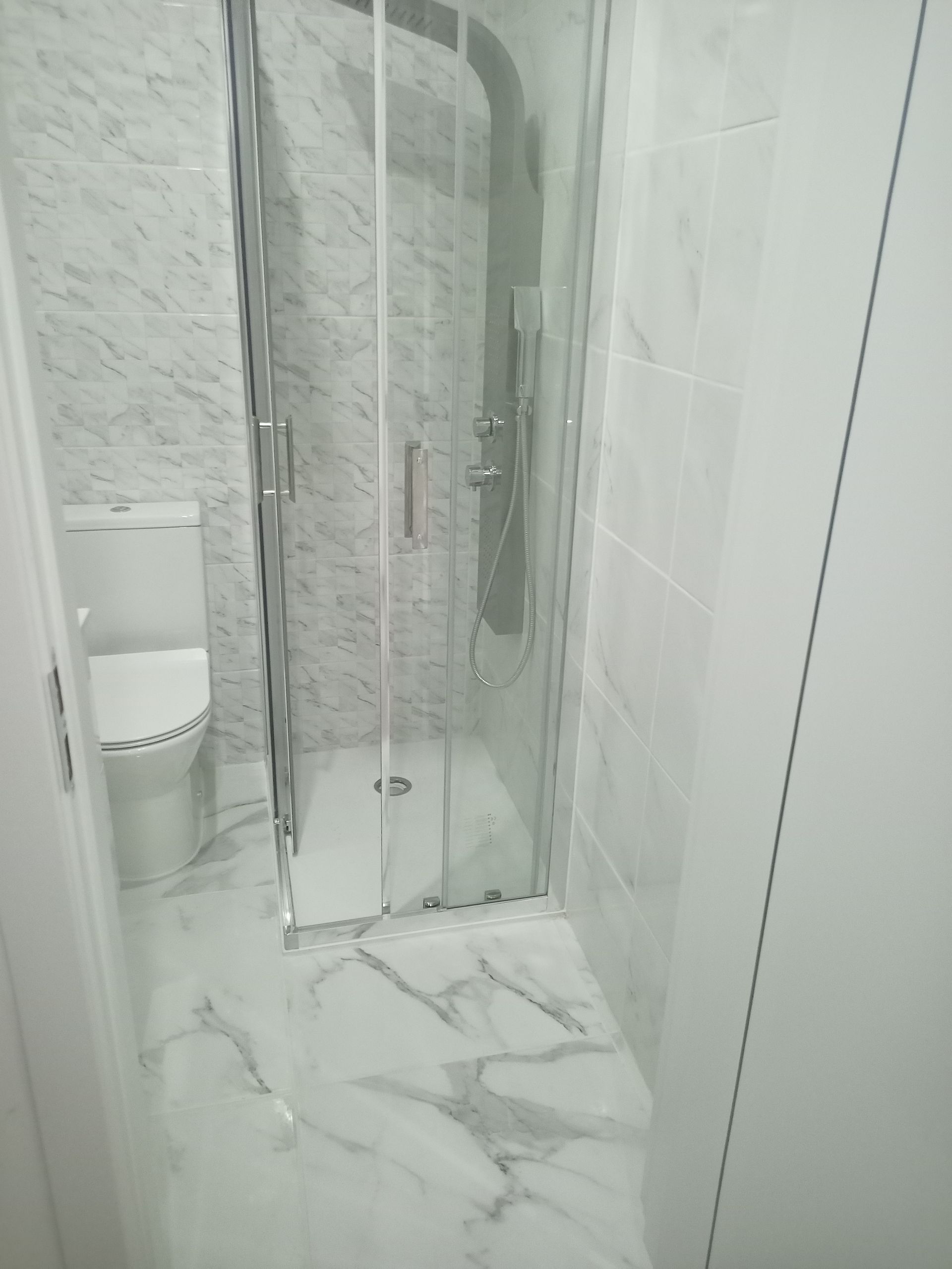 Banheiro com azulejos de mármore branco, box de vidro e vaso sanitário.