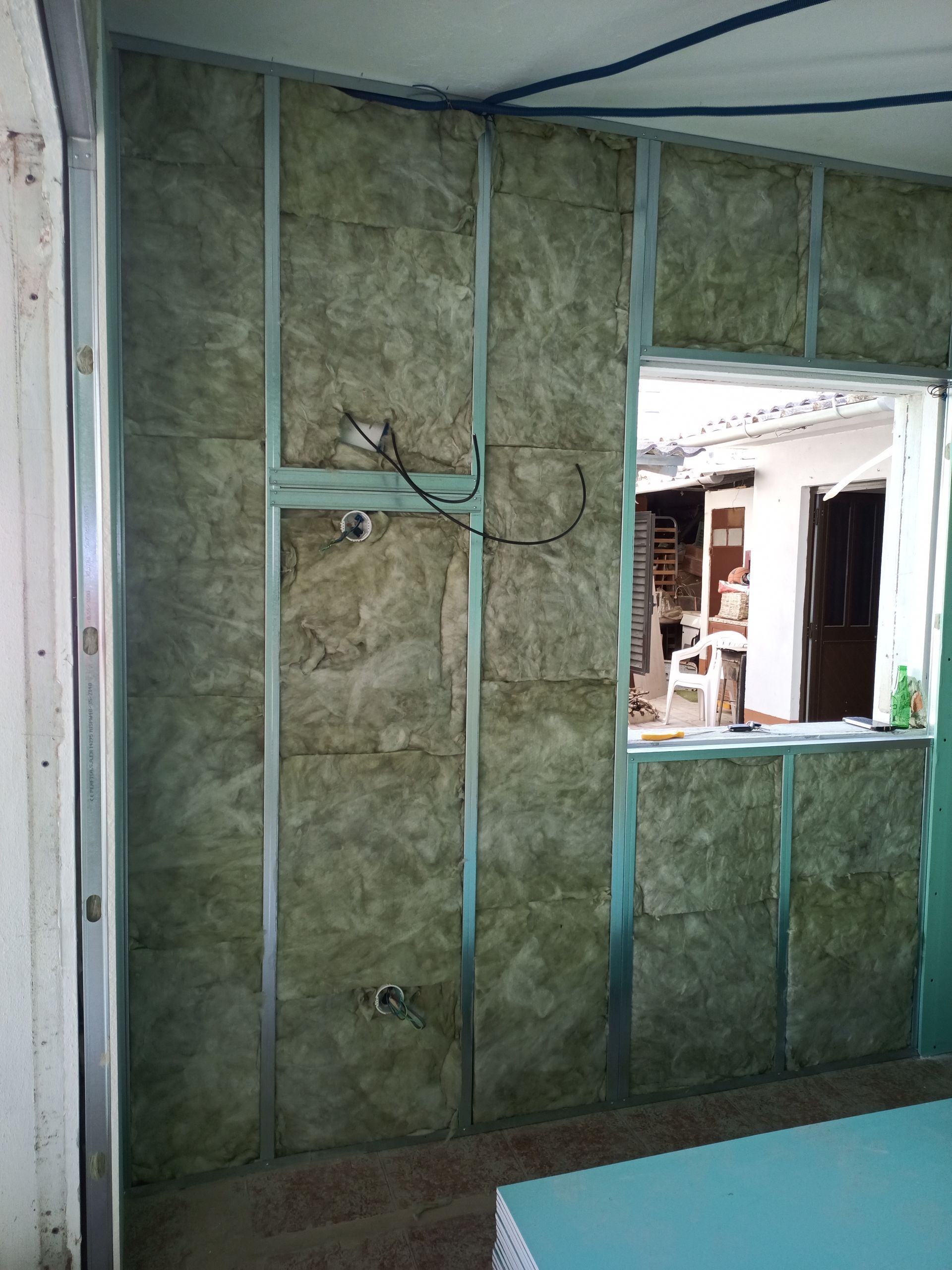 Parede interna em construção com isolamento entre perfis metálicos, abertura de janela e fiação exposta.