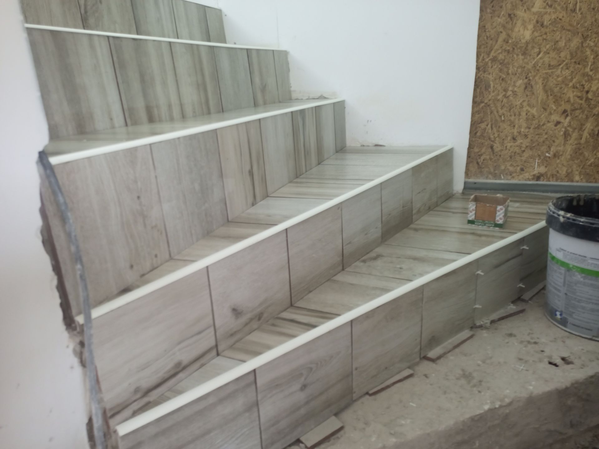 Escada com revestimento em ladrilhos que imitam madeira clara e acabamento branco sendo instalada; paredes e piso inacabados visíveis.