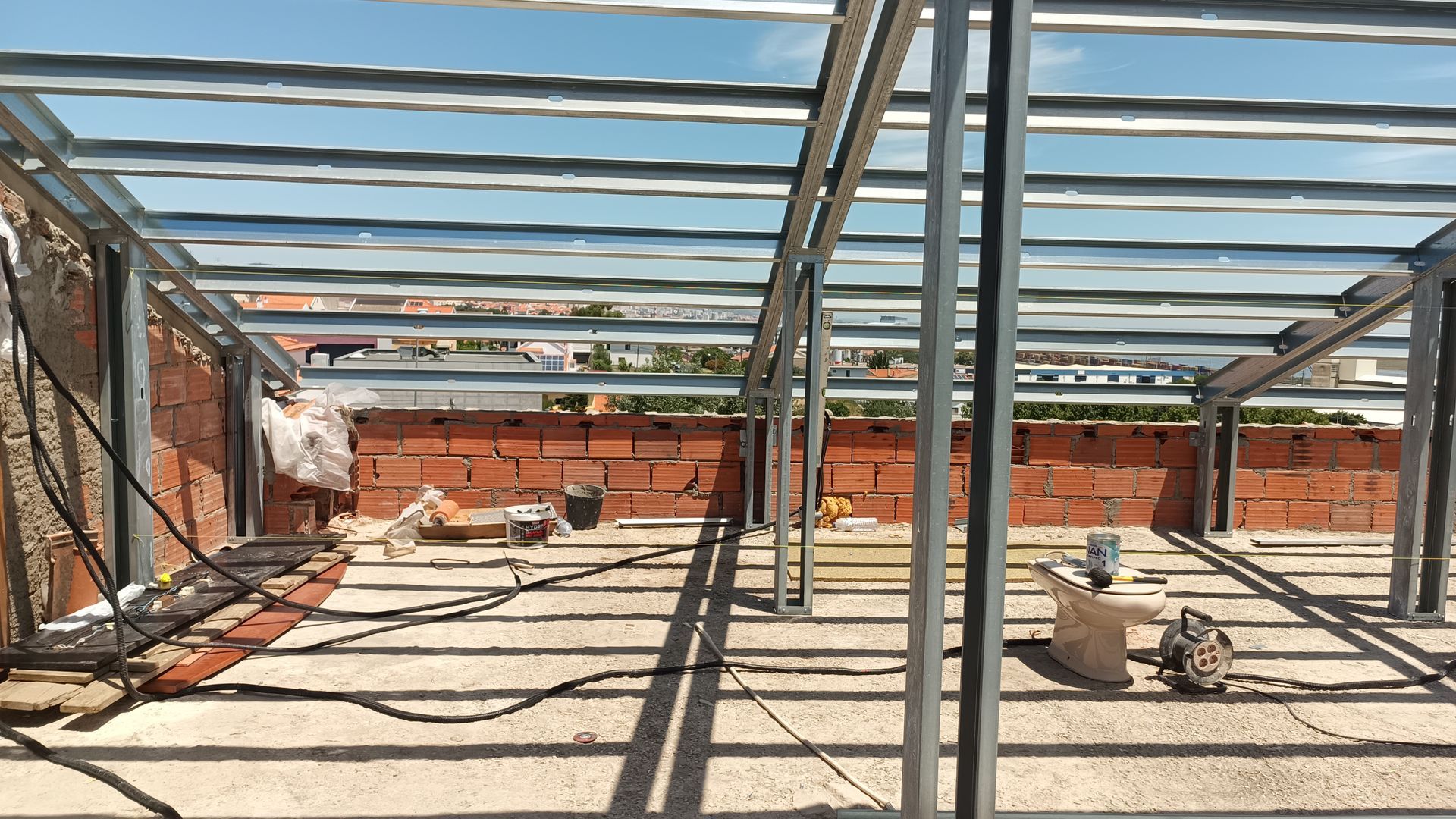 Estrutura metálica de uma construção em andamento; parede de tijolos visível ao fundo.