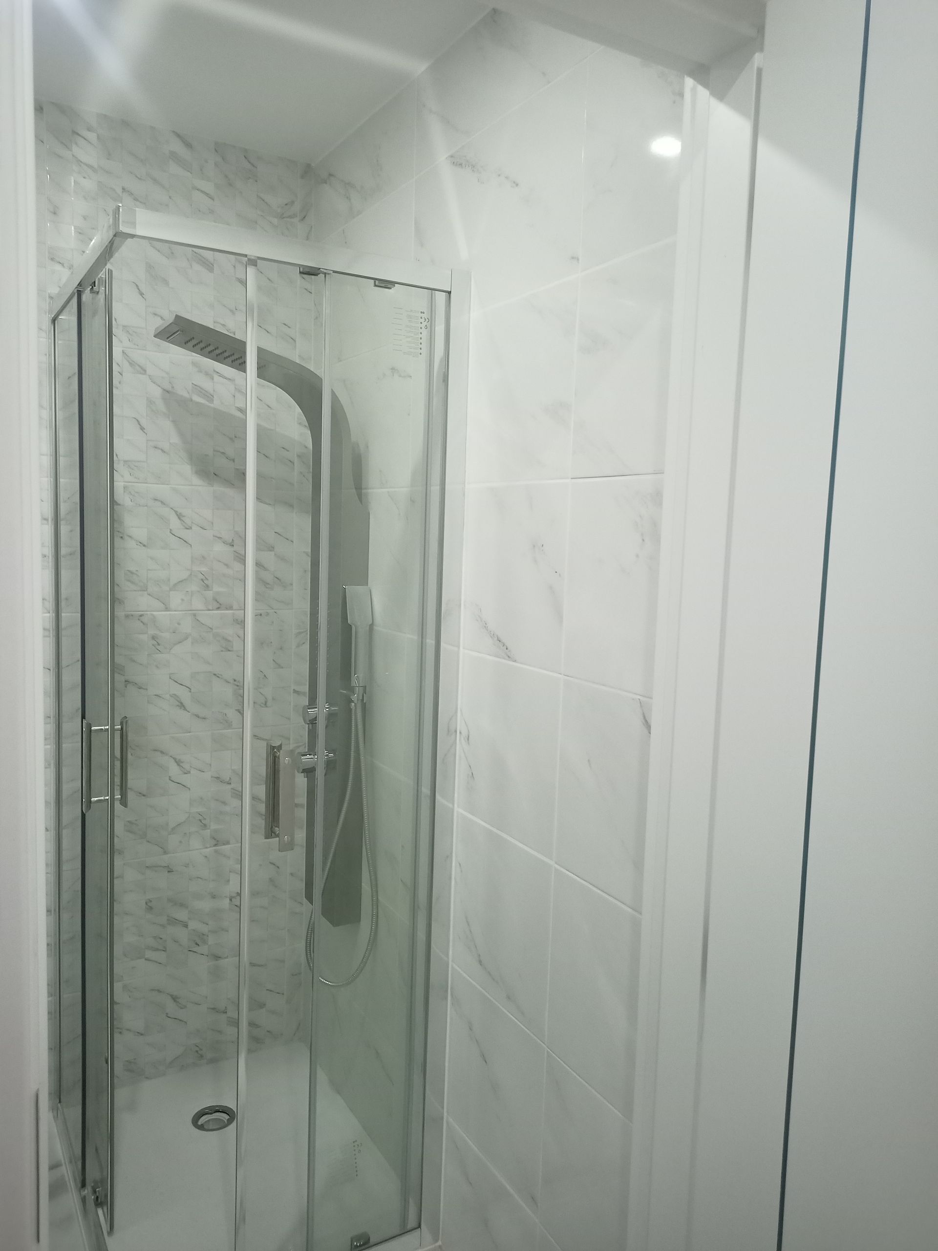 Box de chuveiro em vidro com acessórios prateados em um banheiro com azulejos brancos.