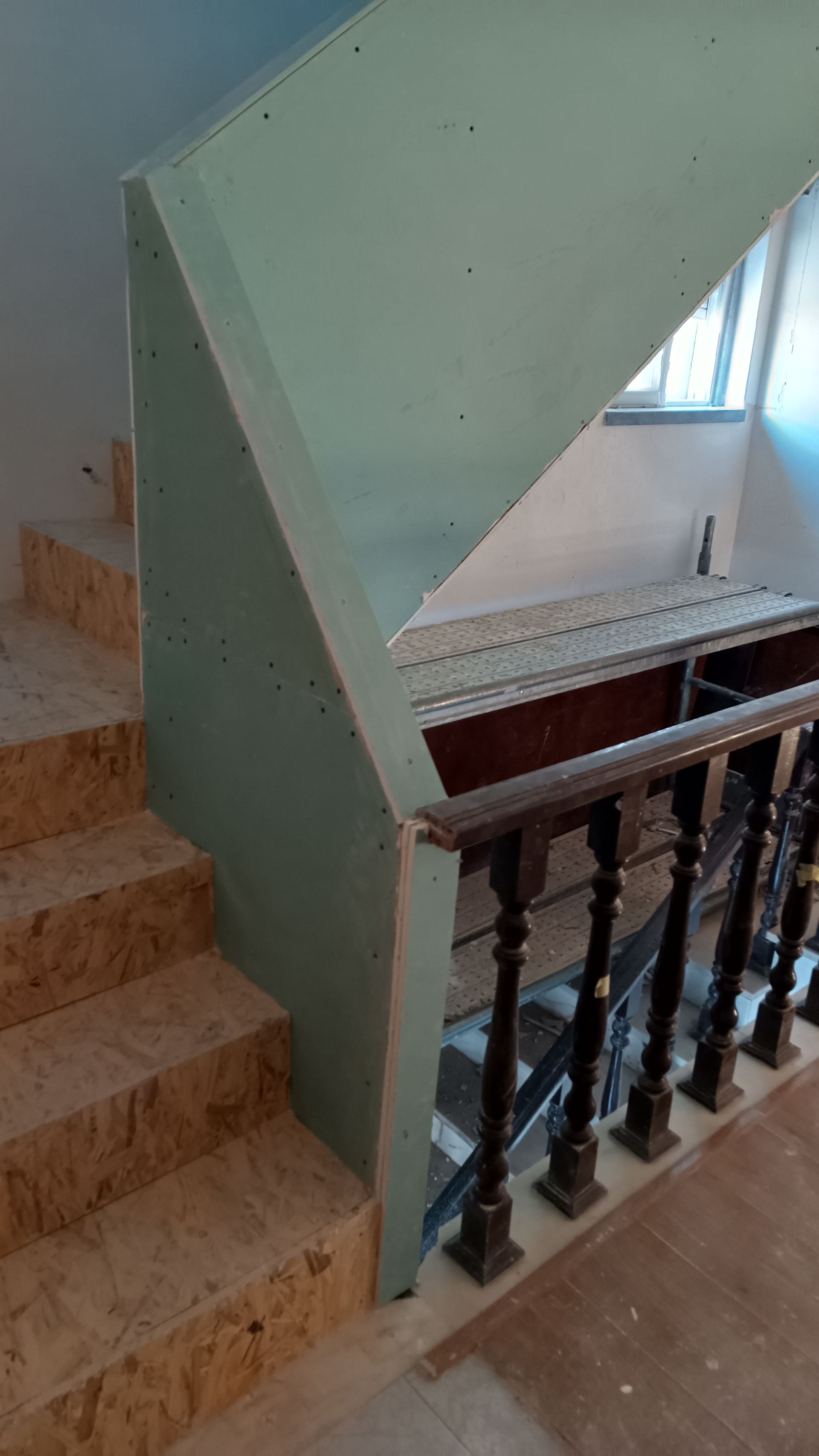 Escada em construção: degraus bege, drywall verde, corrimão de madeira escura e uma janela ao fundo.