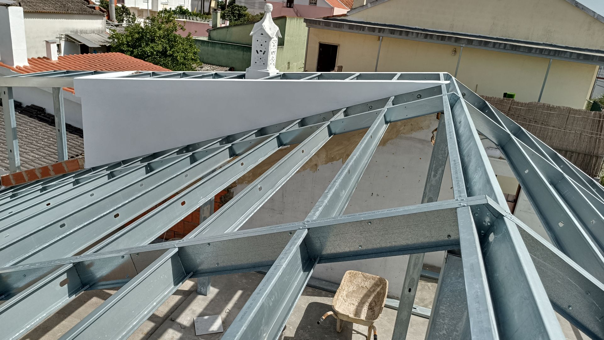 Estrutura metálica do telhado, ambiente externo. Vigas e suportes cinza; prédio branco ao fundo.