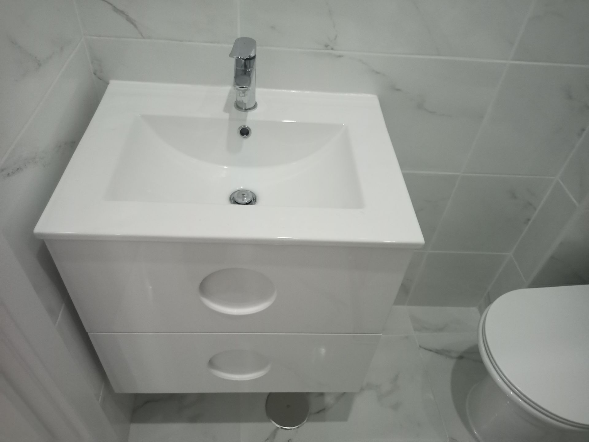 Móvel de banheiro branco suspenso com pia e duas gavetas, torneira cromada e vaso sanitário.