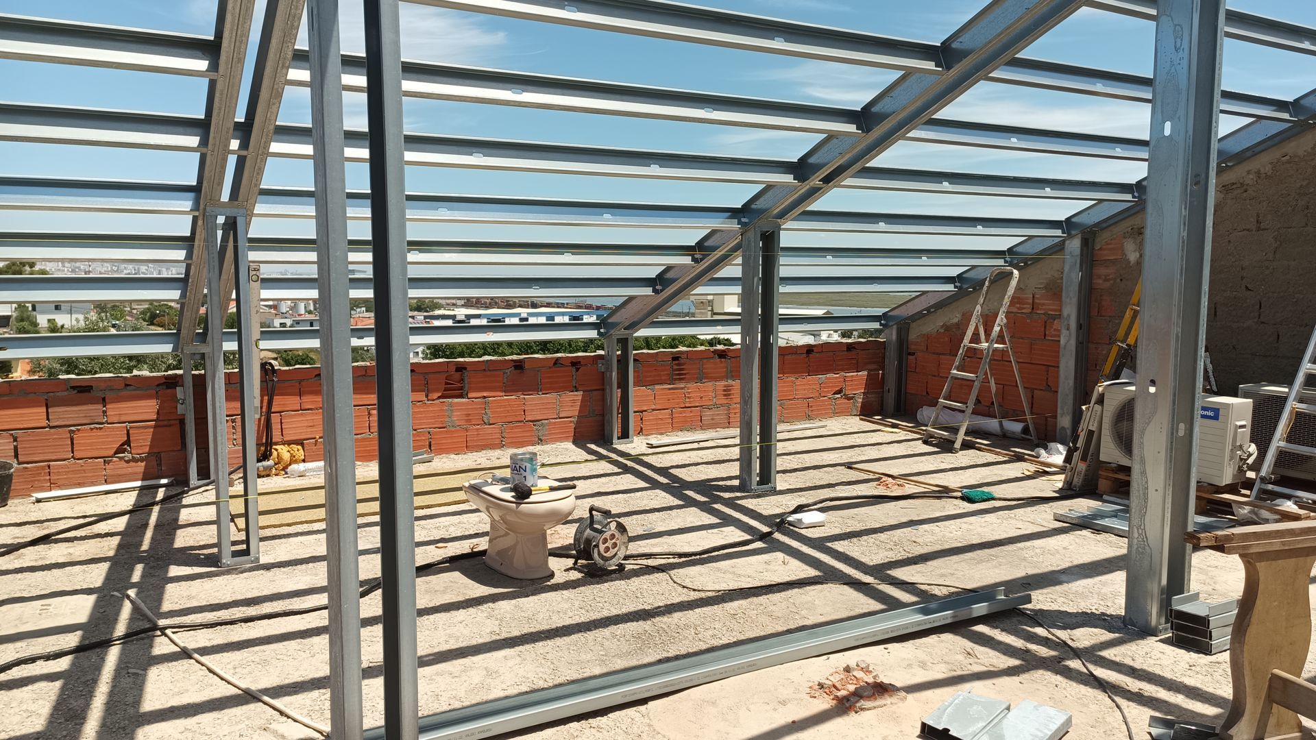 Construção de uma estrutura de telhado com armação metálica sobre uma área com paredes de tijolo, com ferramentas e escada presentes.