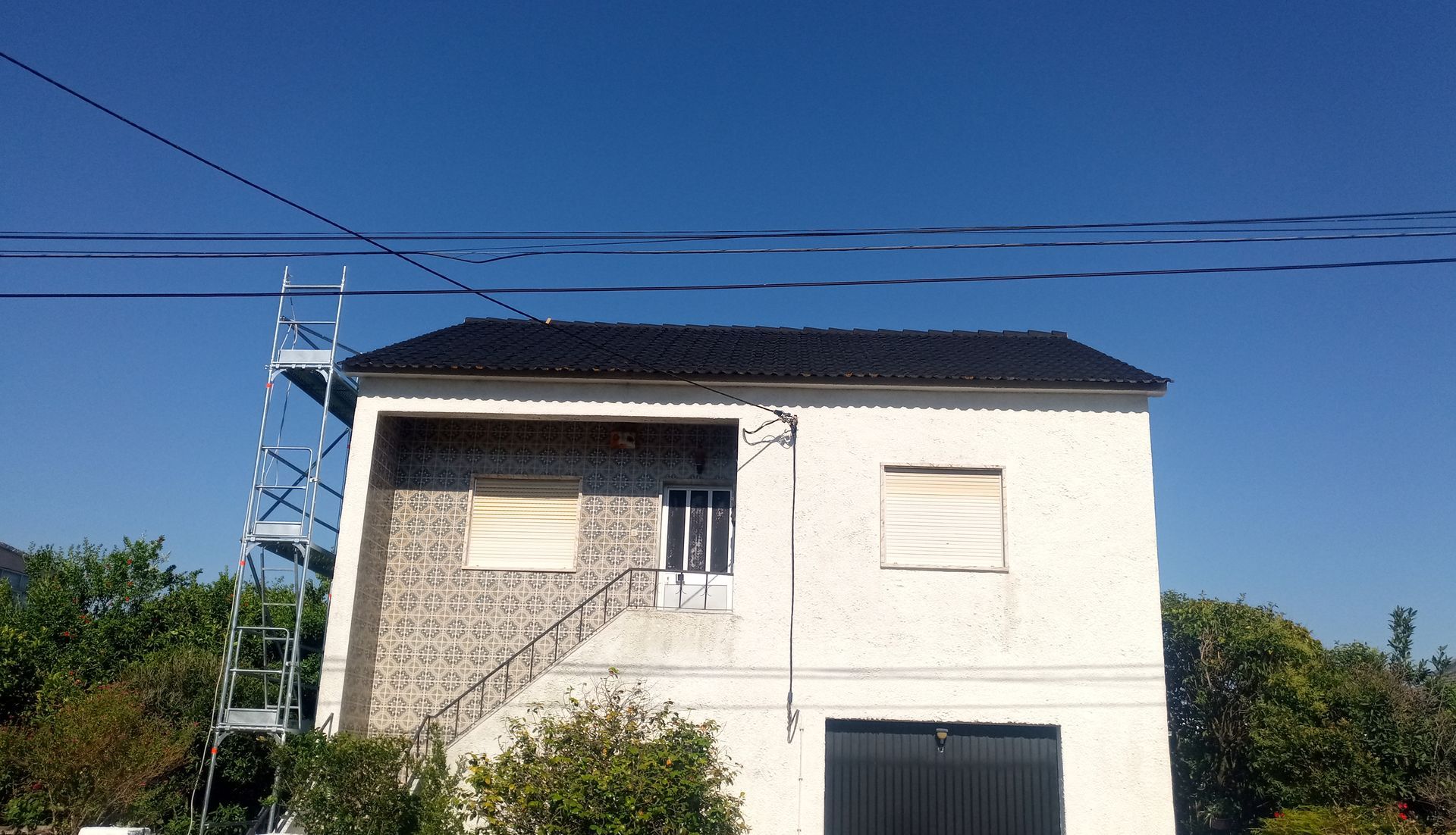 Casa branca de dois andares com telhado preto, andaimes e persianas fechadas contra um céu azul claro.