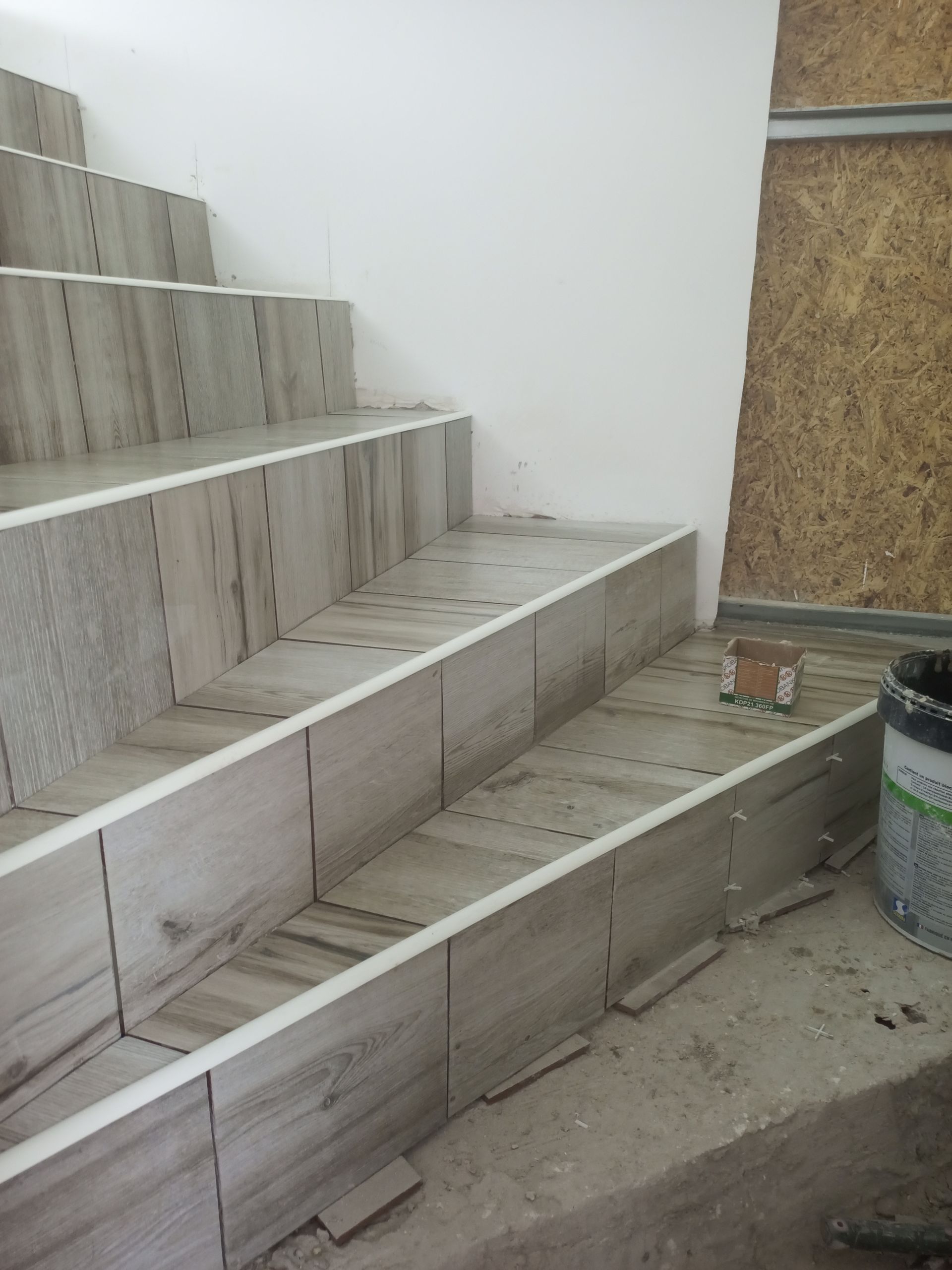 Escada em construção com azulejos claros. Degraus e espelhos parcialmente revestidos com azulejos. Parede branca e painéis marrons presentes.