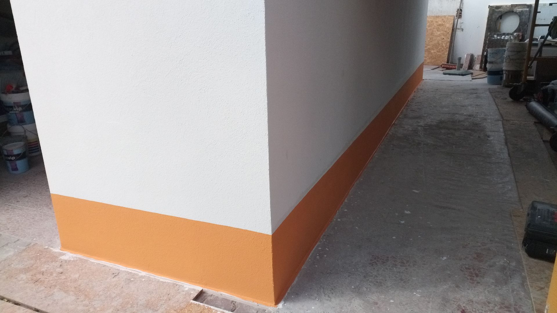 Canto de parede branca com faixa laranja em um ambiente que lembra uma garagem, com ferramentas e suprimentos por perto.