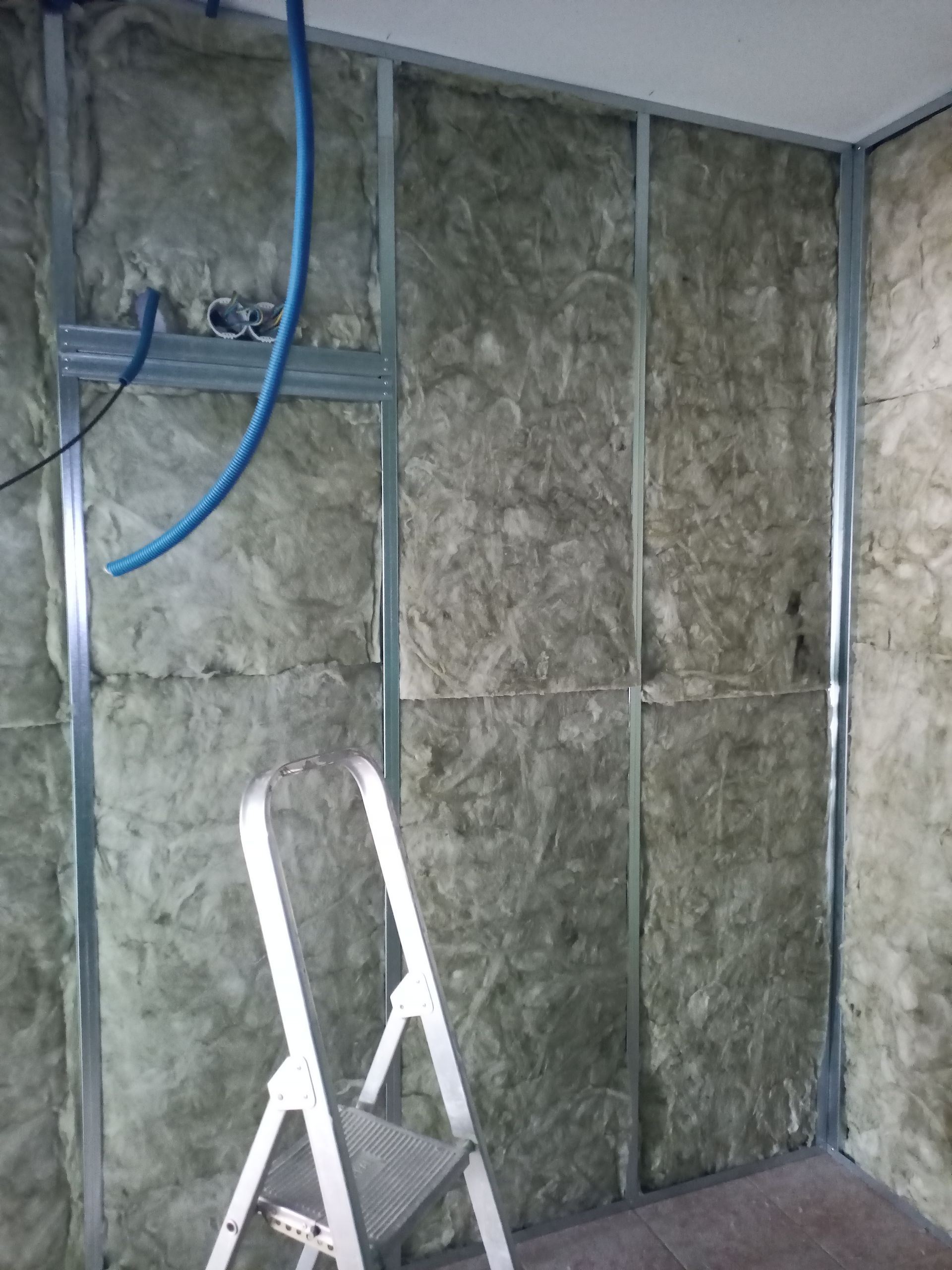 Parede com estrutura metálica em construção, com isolamento térmico, cano azul e uma escada de mão em um cômodo inacabado.