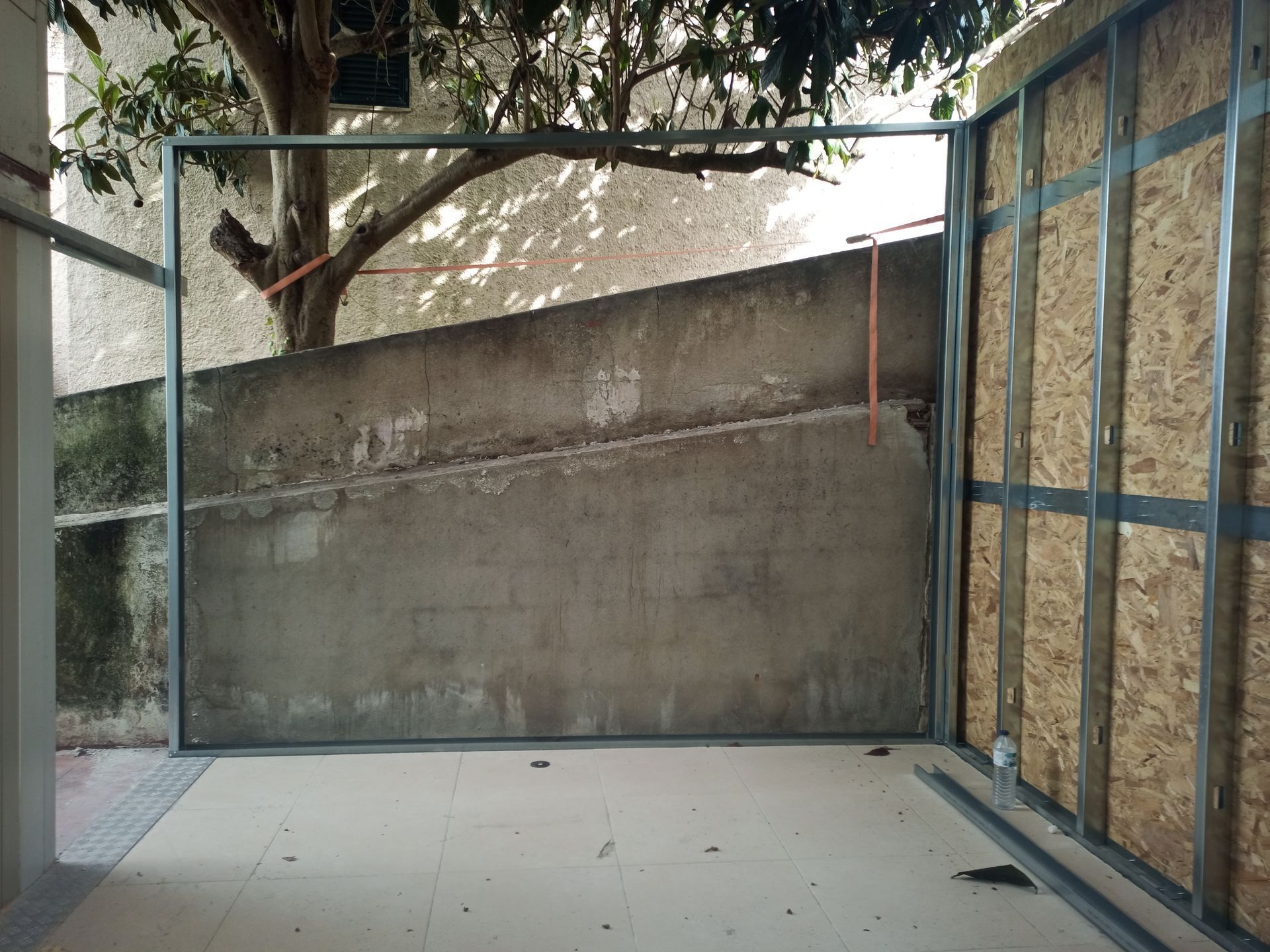 Construção de uma porta em um cômodo, com estrutura de metal, parede de concreto e árvore do lado de fora.