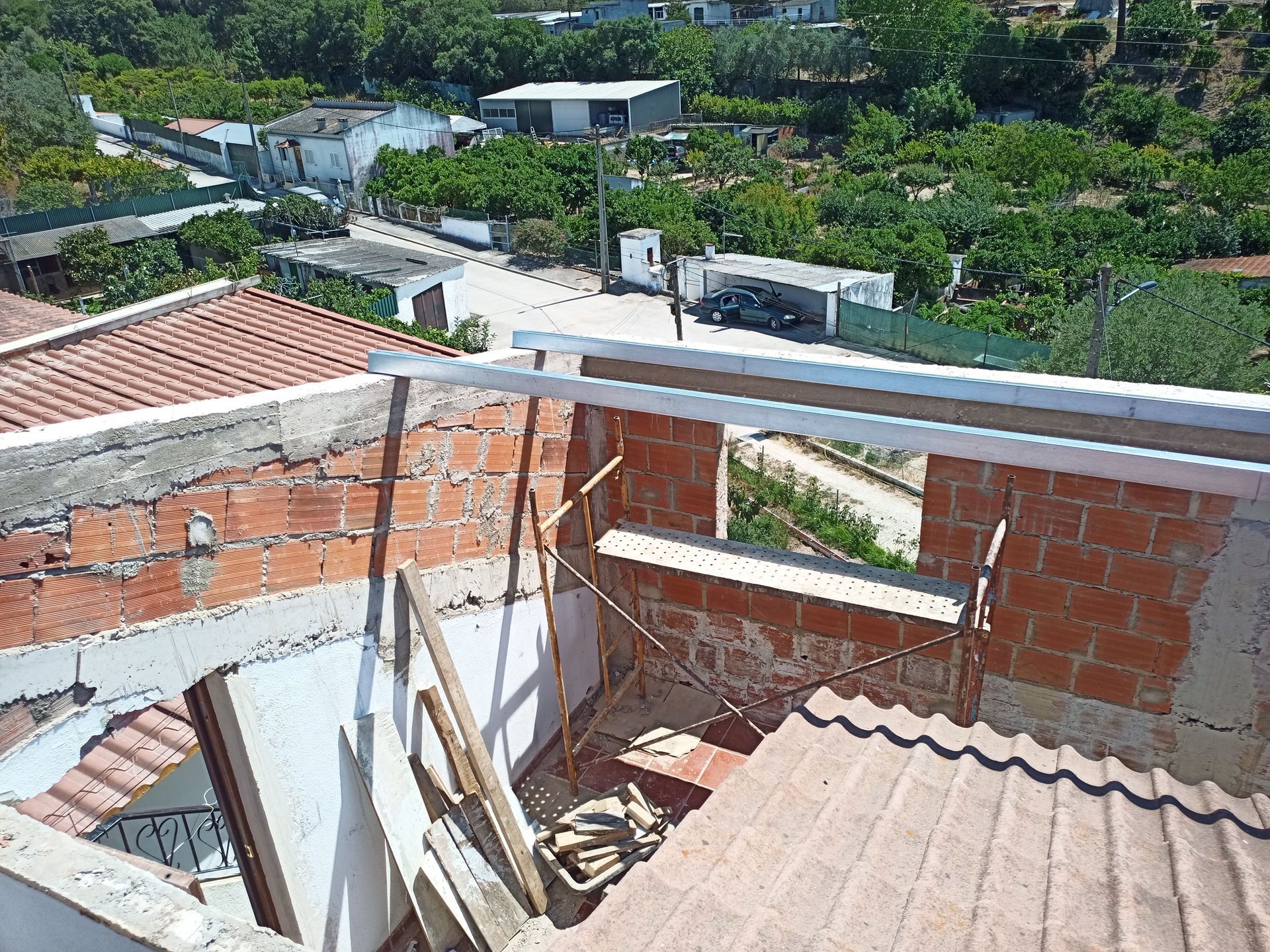 Canteiro de obras com paredes de tijolo vermelho, telhado parcialmente construído e vista para uma área rural com edifícios e árvores.