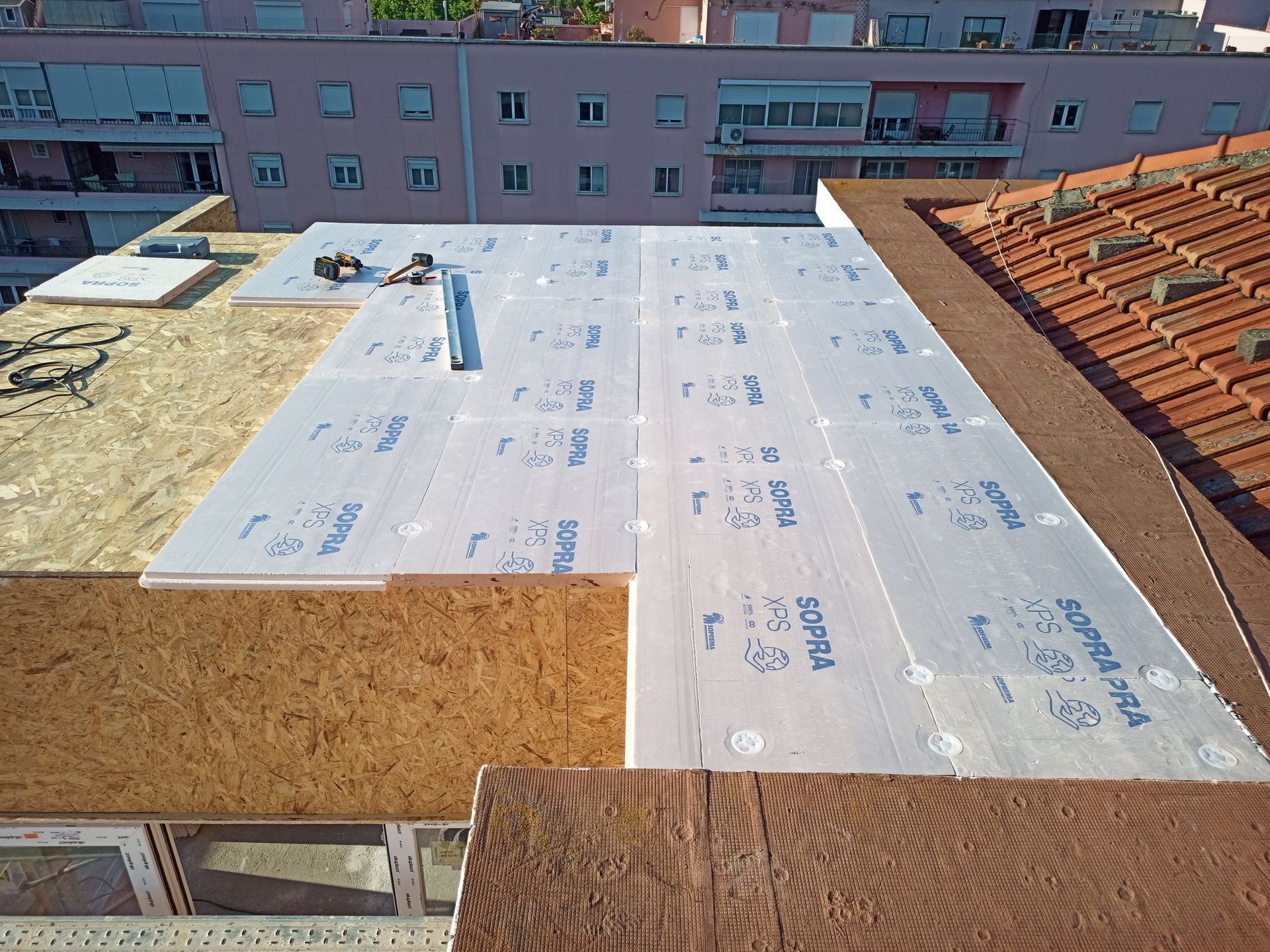 Construção em telhado: Painéis isolantes colocados sobre madeira compensada, ao lado de telhado de telha vermelha, ambiente urbano.