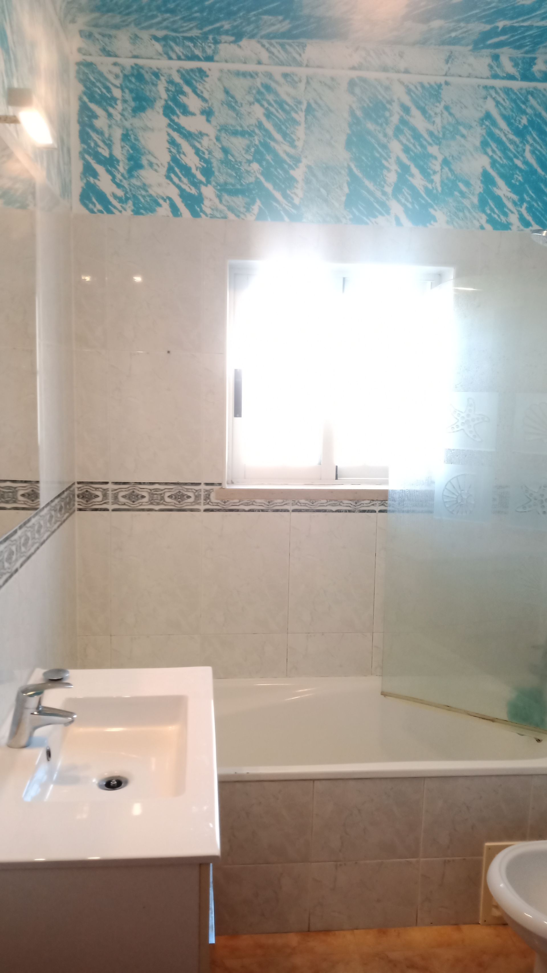 Banheiro com azulejos brancos e azuis, pia, banheira e janela com bastante luz.