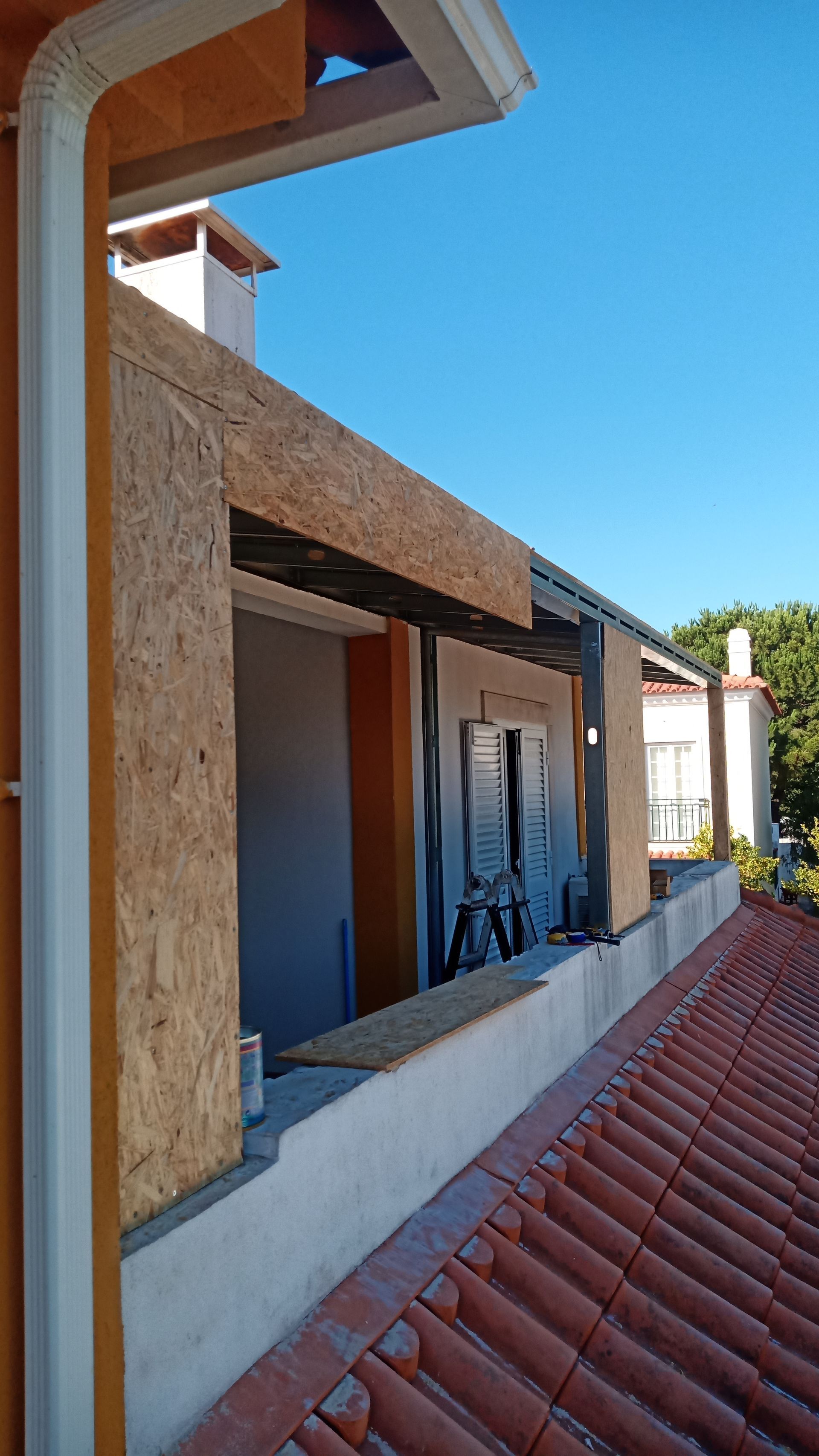 Prédio em construção com estrutura de madeira aparente. Telhado de telha vermelha, calha branca e céu azul.