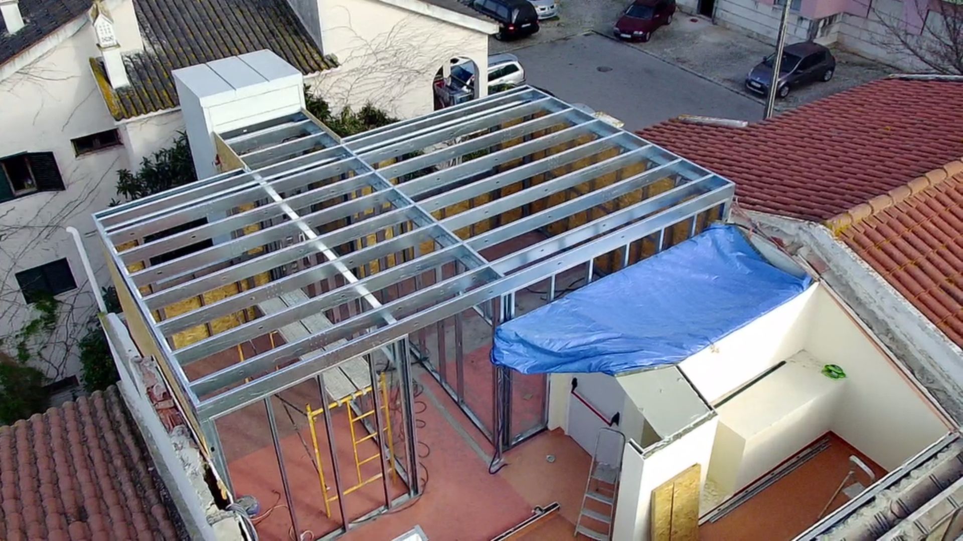 Estrutura metálica sobre um telhado, coberta com uma lona azul, com vista para uma rua com carros e prédios.