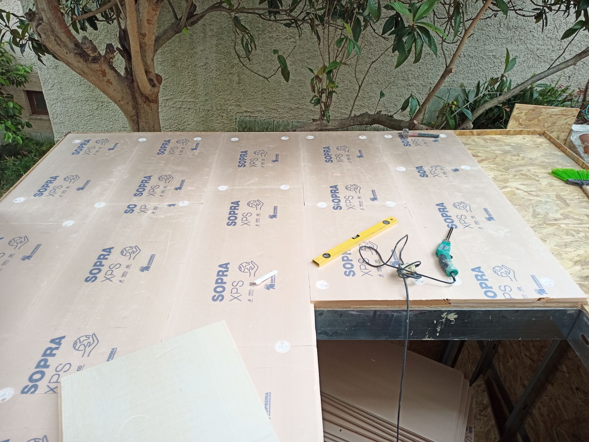 Construção de um telhado plano, com placas de isolamento e ferramentas (nível, furadeira) dispostas. Ao ar livre, perto de uma árvore.