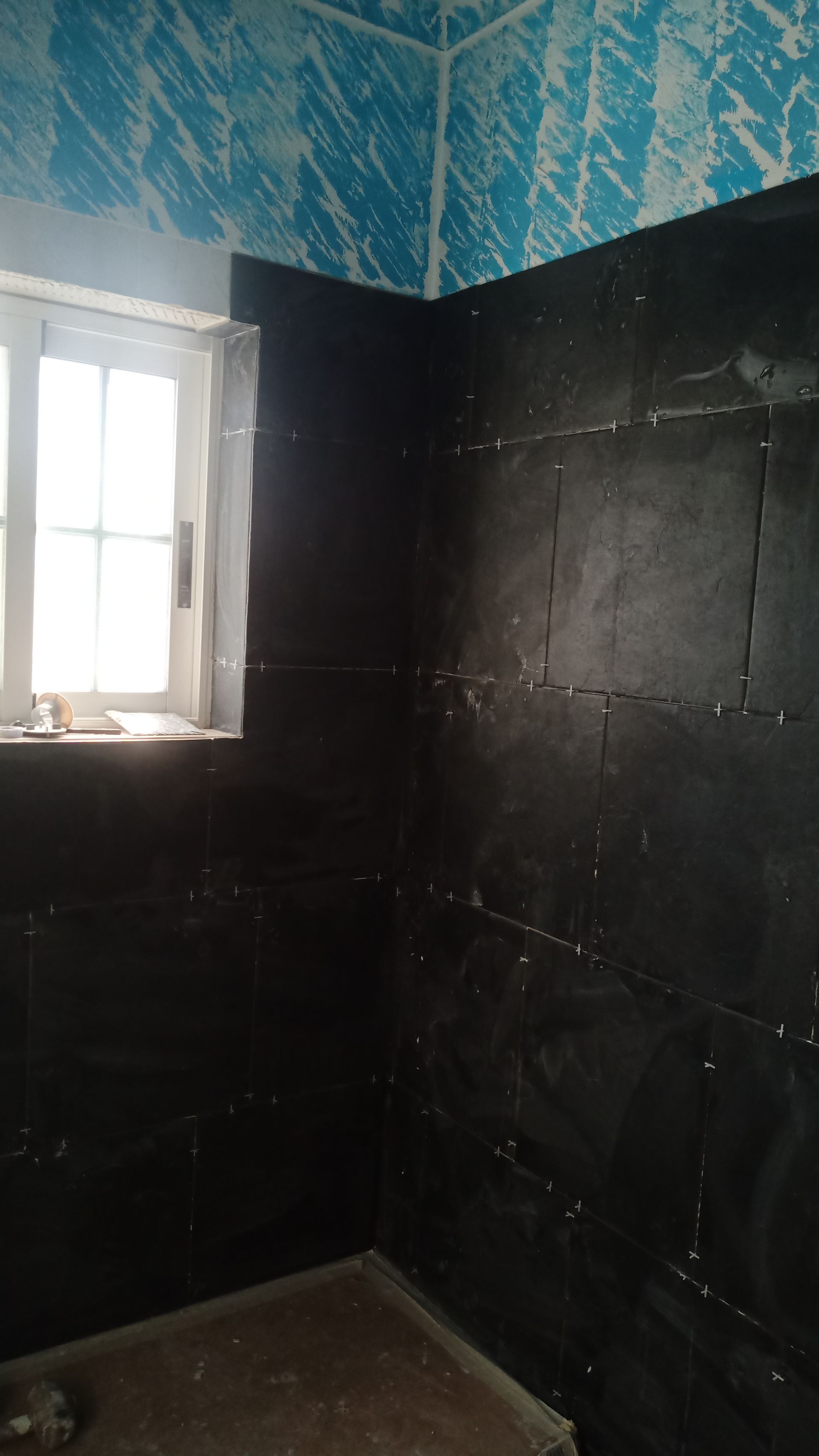 Canto do banheiro com azulejos pretos nas paredes, papel de parede estampado azul e uma janela.