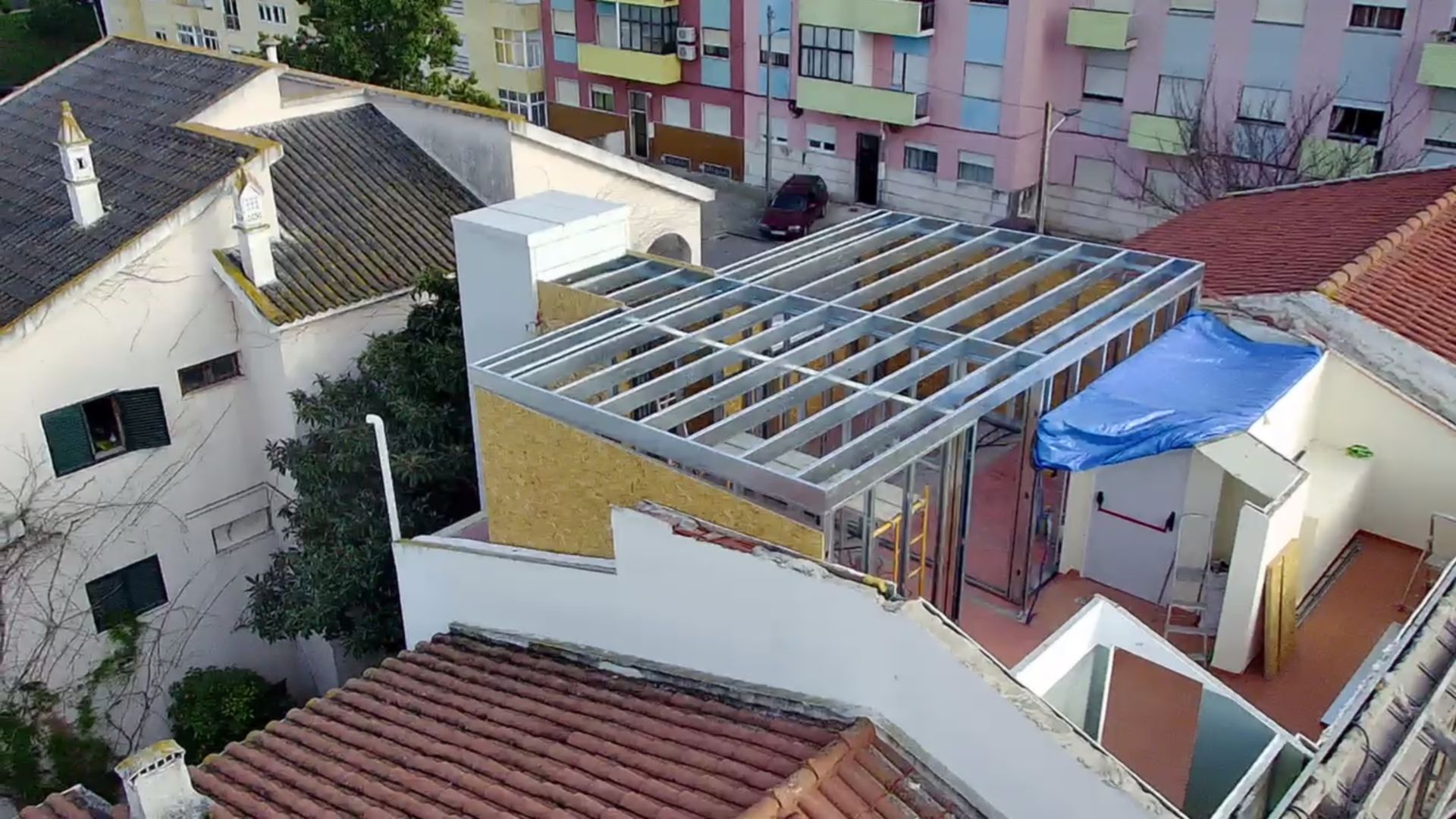Construção de um edifício moderno com estrutura metálica e paredes de tijolo amarelo, rodeado por casas mais antigas com telhados de telha vermelha.