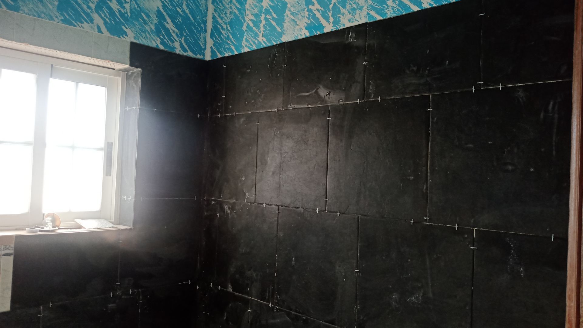 Interior do banheiro: Azulejos pretos instalados na parede, papel de parede azul e branco acima, janela à esquerda.