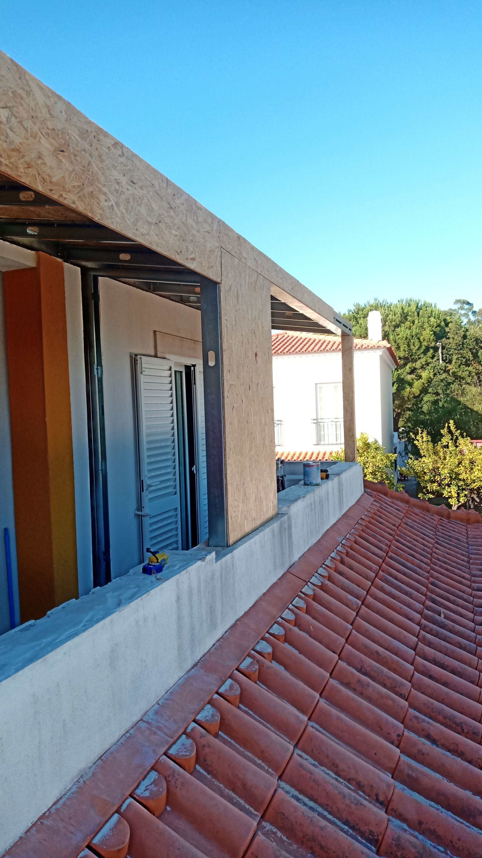 Vista do terraço: canteiro de obras com estrutura aparente, beiral de concreto e telhado de telha vermelha contra um céu azul.
