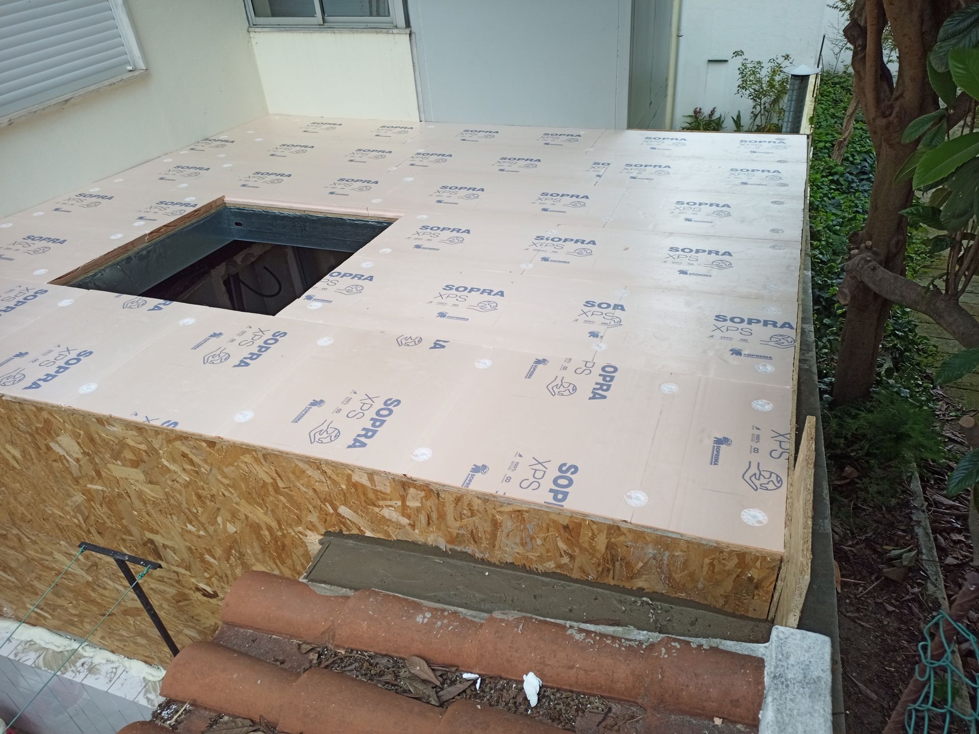 Uma estrutura retangular de madeira com painéis isolantes na parte superior, abertura quadrada, rodeada por elementos exteriores.