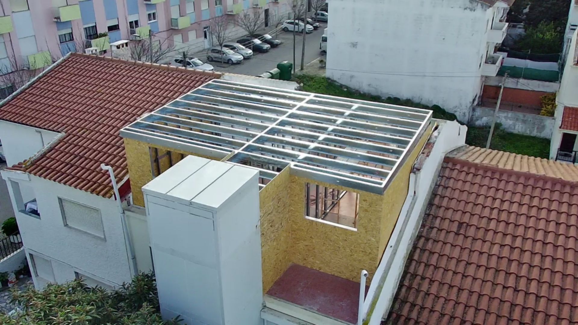 Uma vista aérea mostra um edifício em construção, com o telhado parcialmente concluído, e edifícios residenciais ao redor.