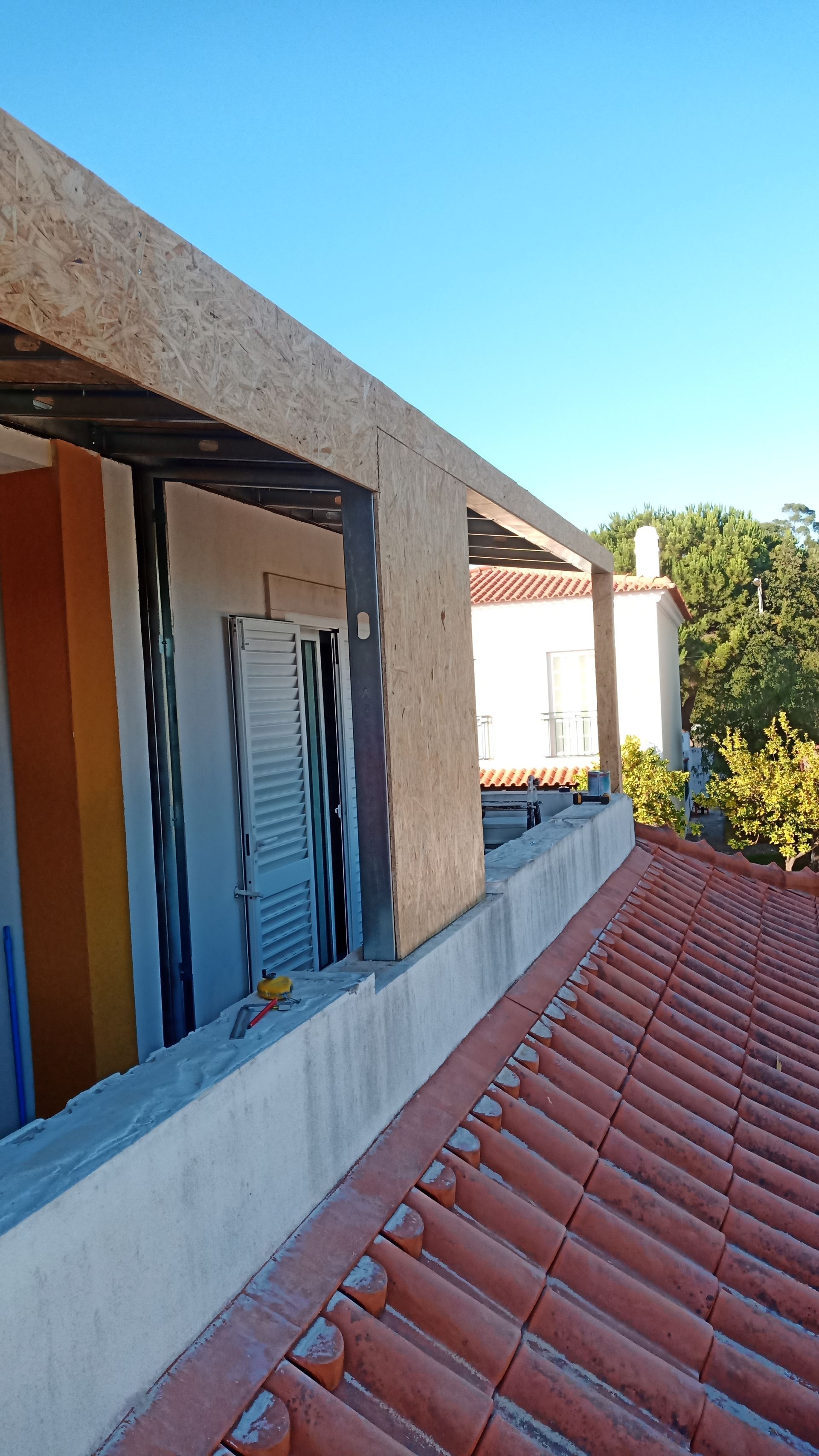 Construção de um edifício com telhado de telhas vermelhas, uma parede inacabada com uma janela e um céu azul claro.