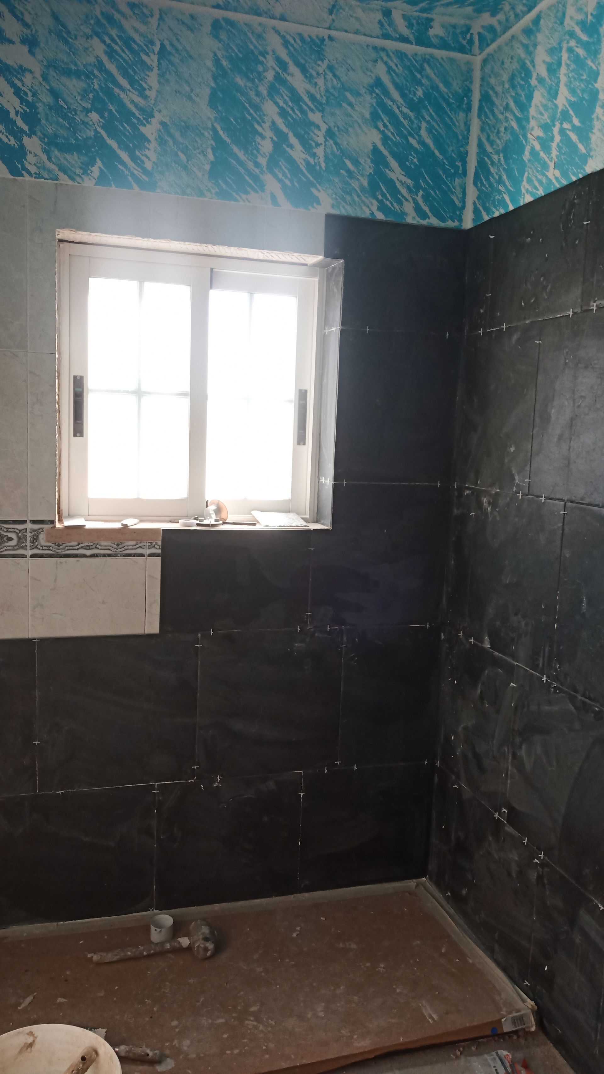 Banheiro com instalação de azulejos inacabada; azulejos pretos na parede, janela e azulejos estampados azuis.