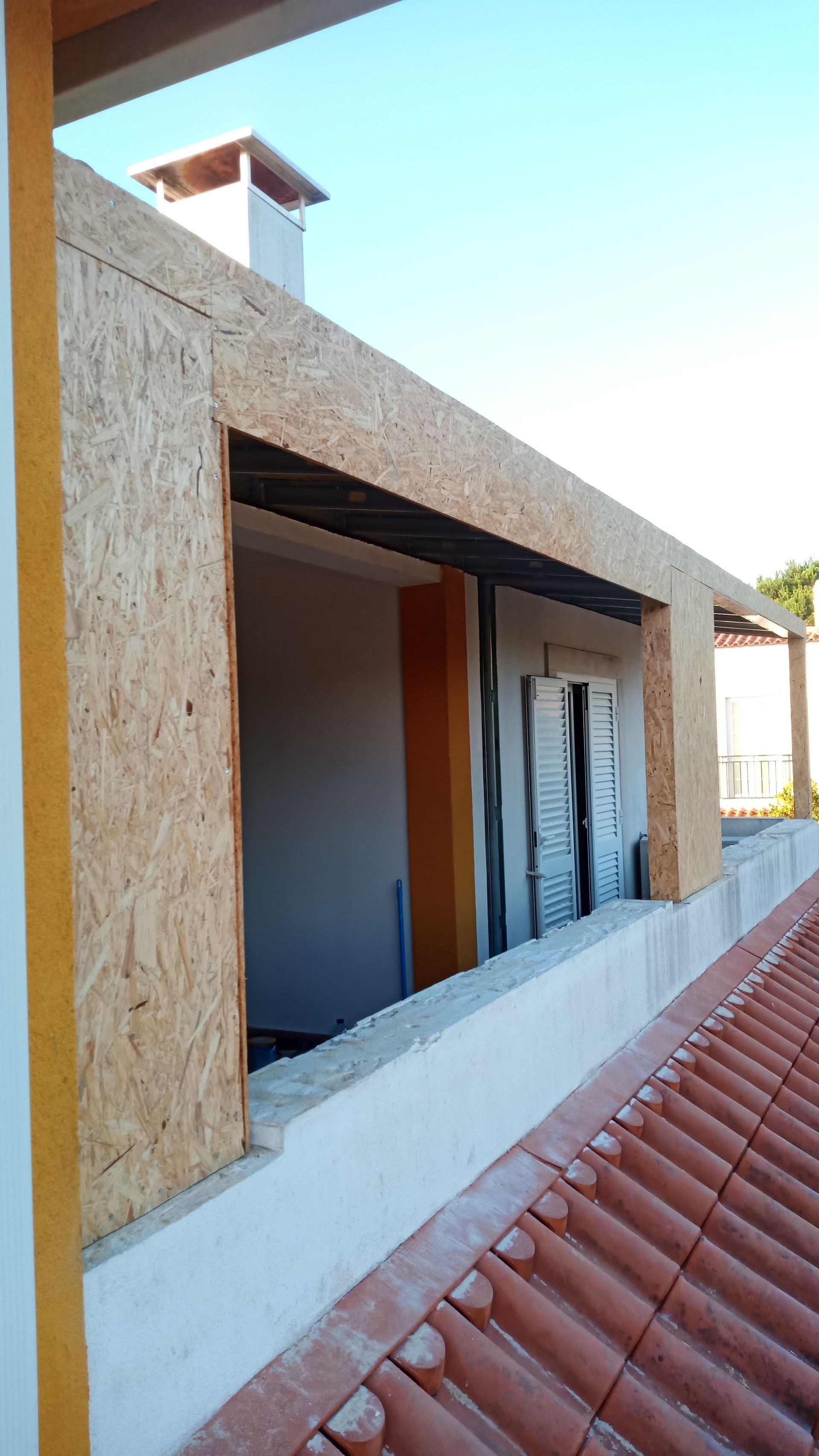 Construção de uma moldura de janela com painéis OSB e peitoril de concreto, tendo como pano de fundo um telhado de telha vermelha e um céu azul.
