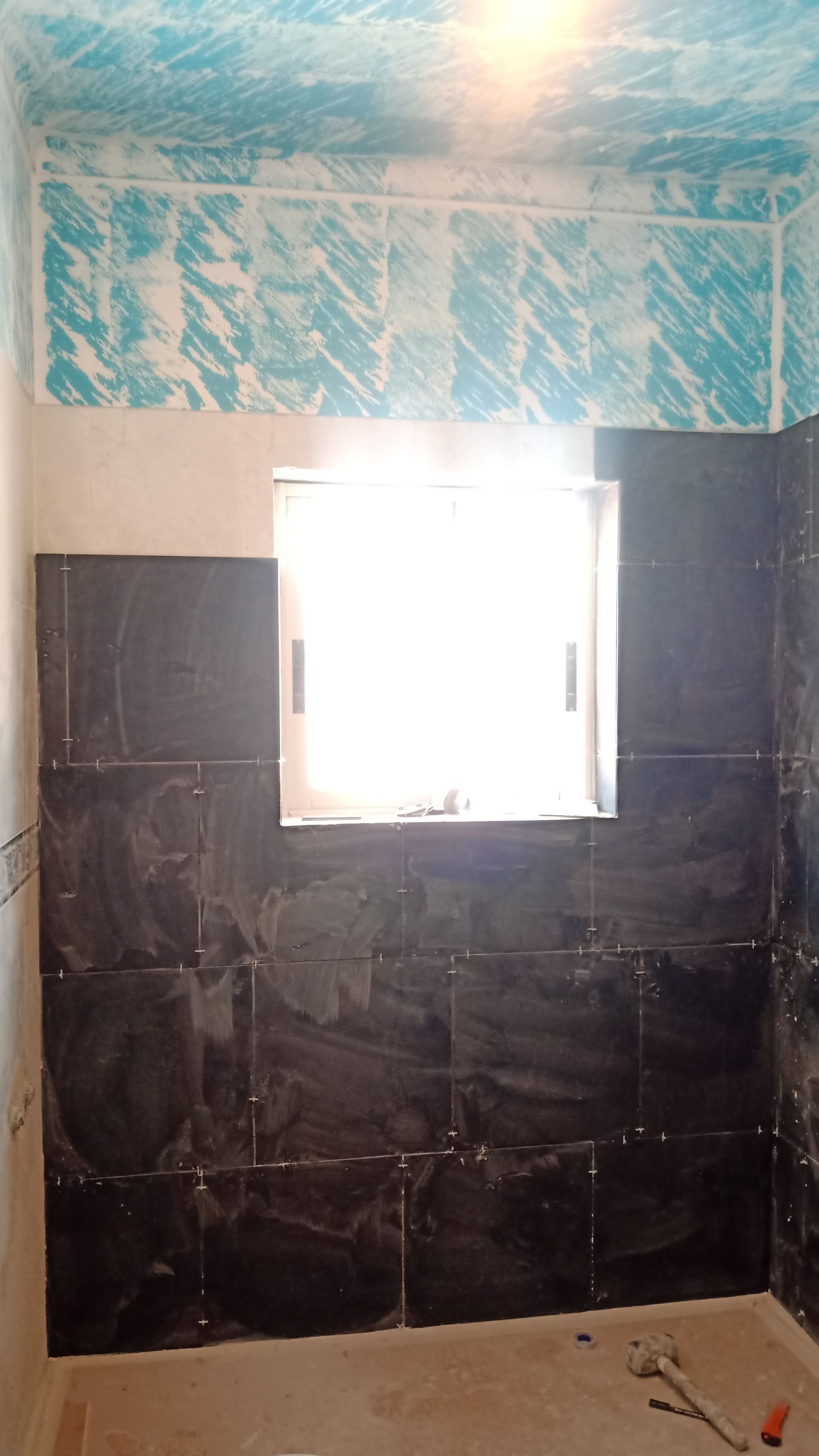 Banheiro com paredes revestidas de azulejos escuros, uma janela e teto pintado de azul.