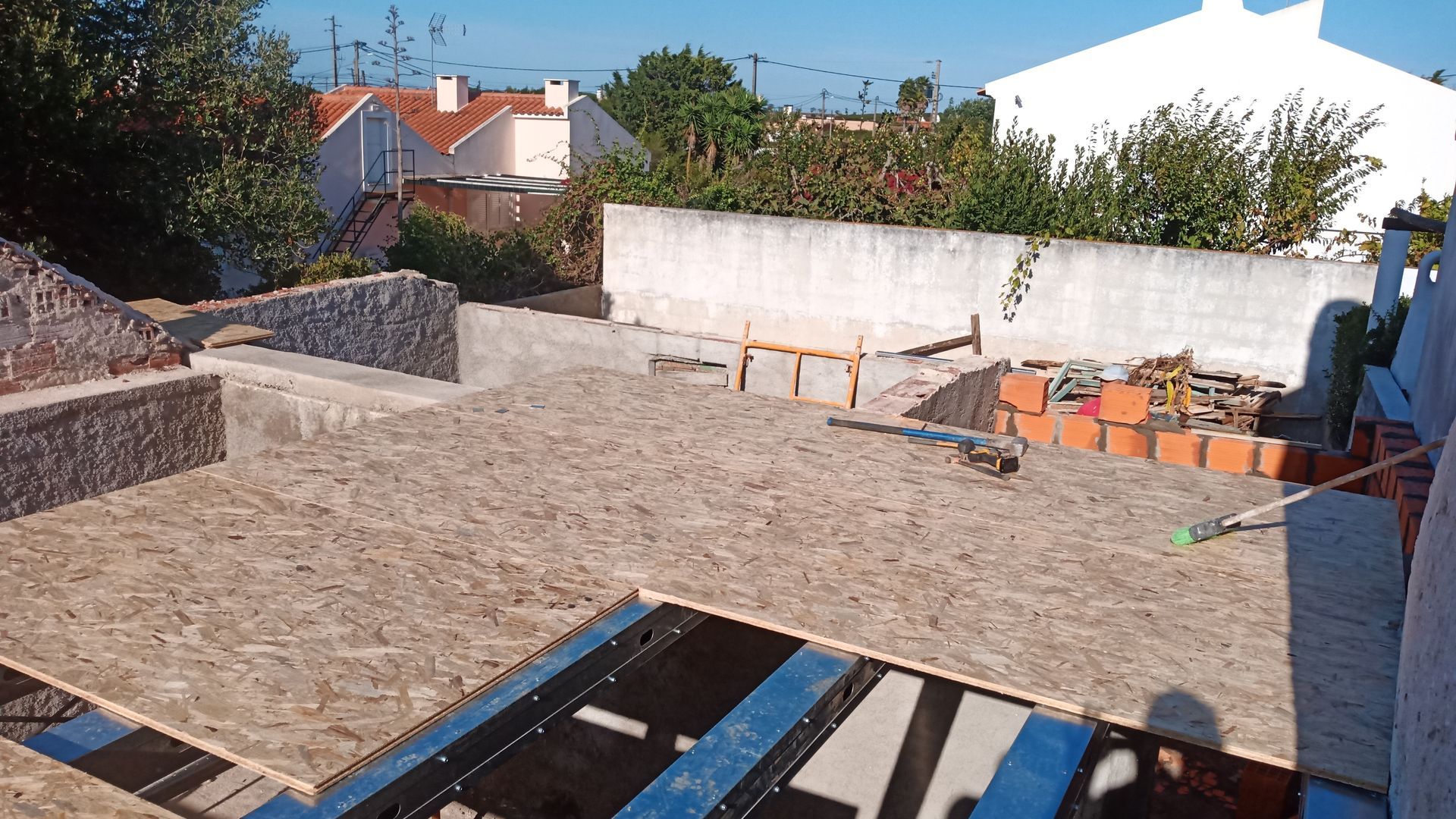 Canteiro de obras com painéis de madeira e materiais de construção. Casas e árvores ao fundo. Luz solar intensa.