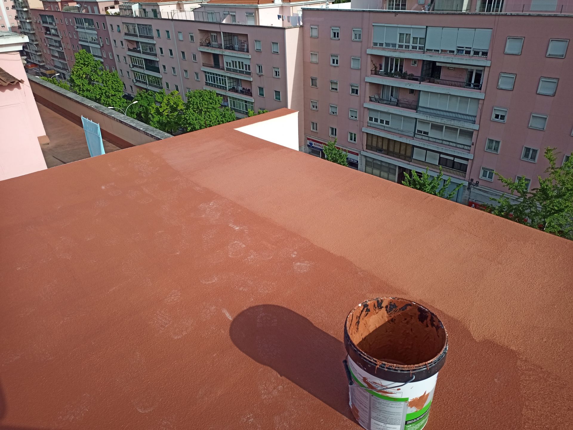 Vista de um telhado: um balde de tinta marrom repousa sobre uma superfície vermelha recém-pintada; prédios de apartamentos ao fundo.