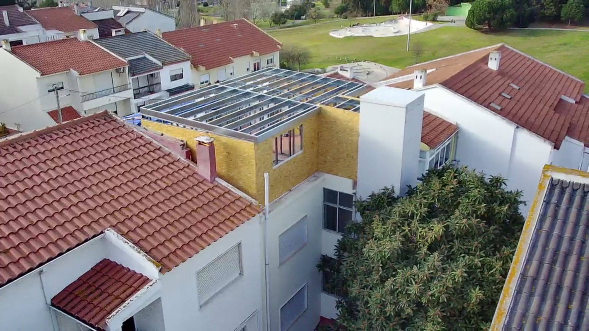 Painéis solares no telhado de um prédio com fachada em terracota e branca, árvores verdes e gramado verde ao fundo.