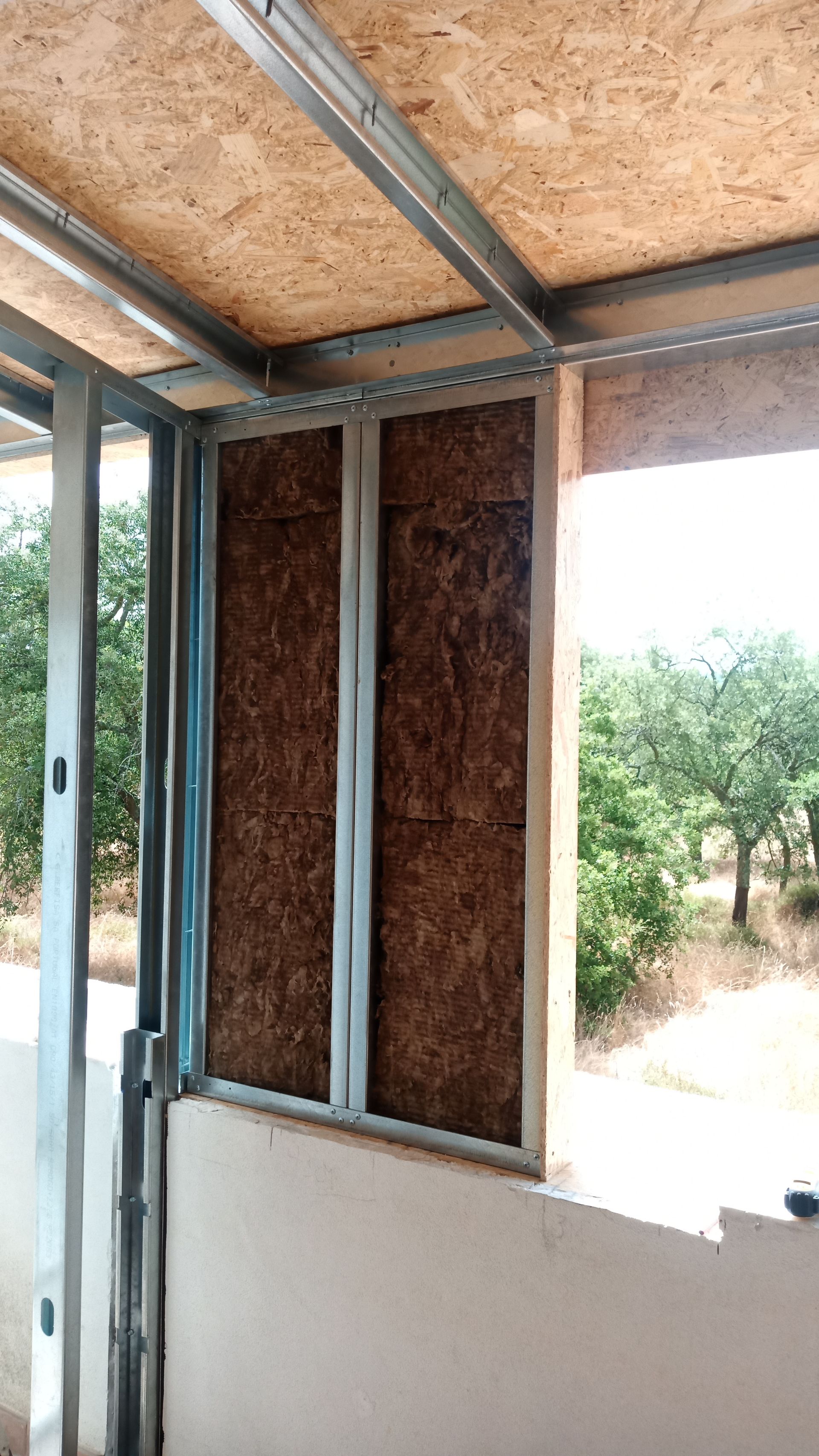 Construção da parede interna: estrutura metálica com painéis de madeira e isolamento aparente, próximo a uma janela aberta.