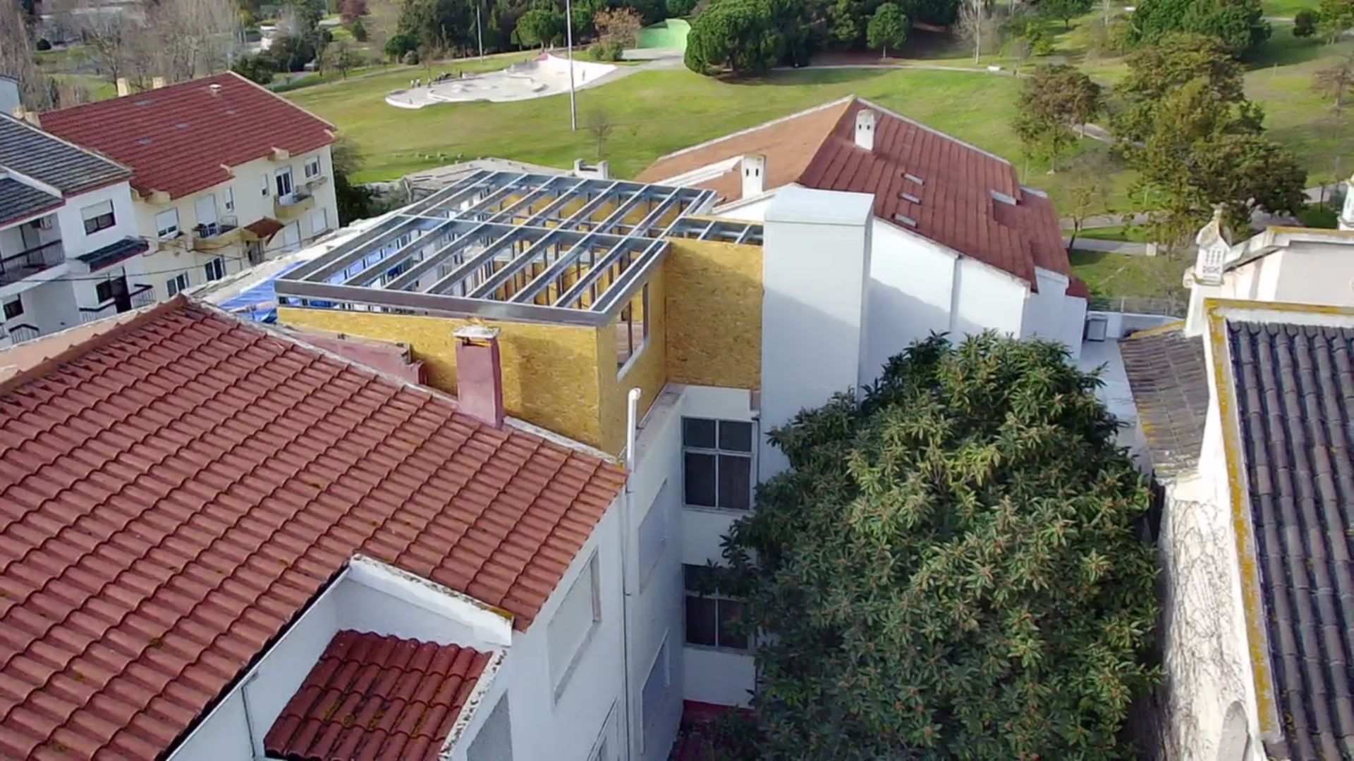 Edifícios com telhados de telha vermelha; um deles com a estrutura metálica do telhado exposta. Árvores verdes e uma área gramada são visíveis.
