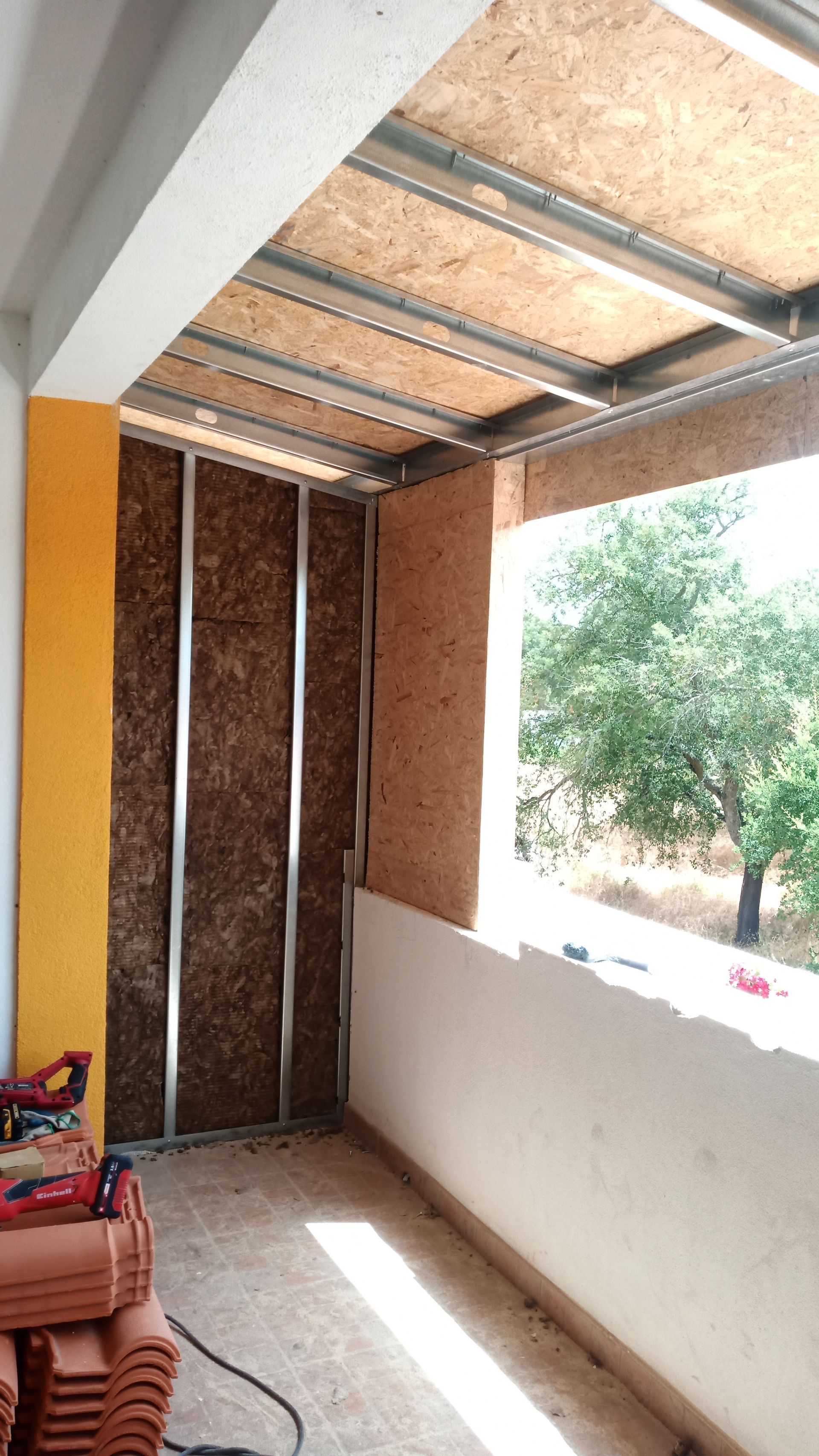 Varanda em construção. Estrutura metálica e isolamento de madeira, com vista para a vegetação externa.