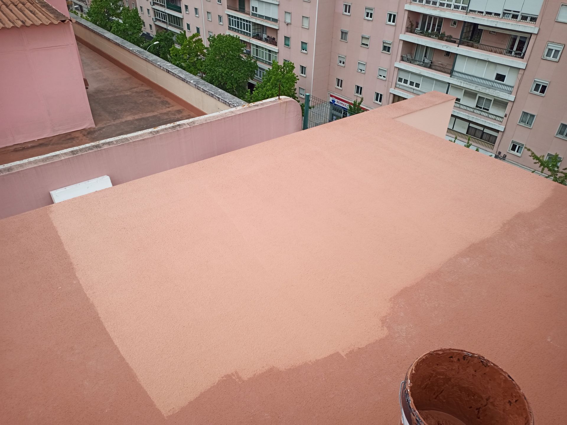 Telhado sendo pintado com revestimento impermeabilizante marrom-avermelhado; um balde está próximo.