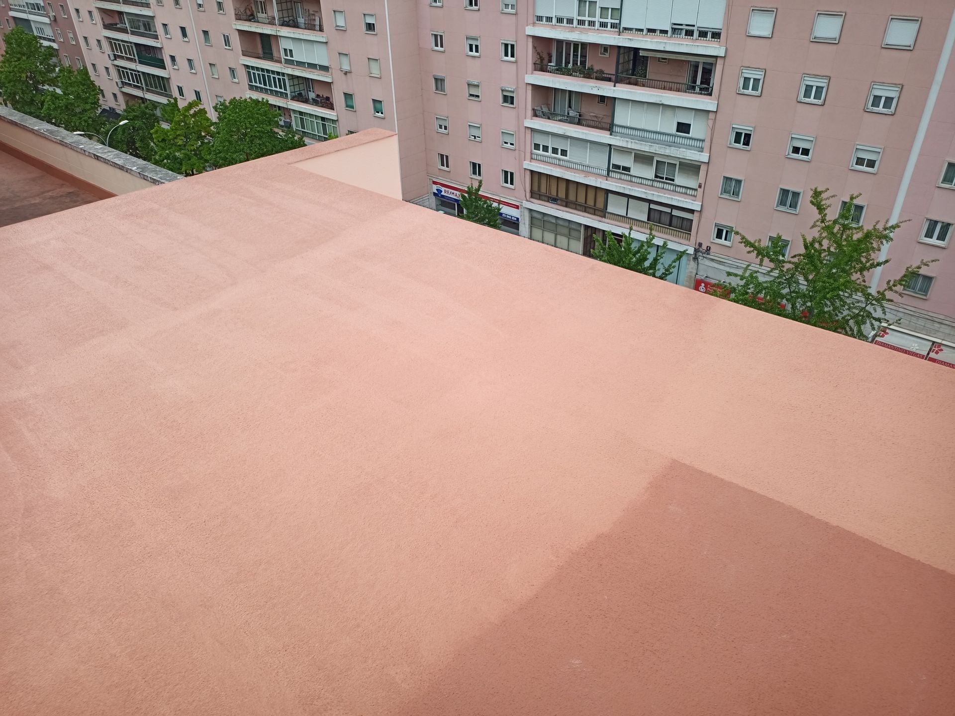 Vista de um telhado plano e avermelhado com prédios de apartamentos ao fundo. Árvores verdes e o céu são visíveis.