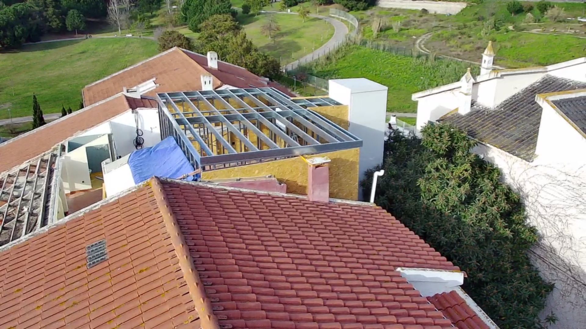 Vista aérea de edifícios com telhados de telha vermelha, um canteiro de obras com estrutura metálica e uma paisagem verde.