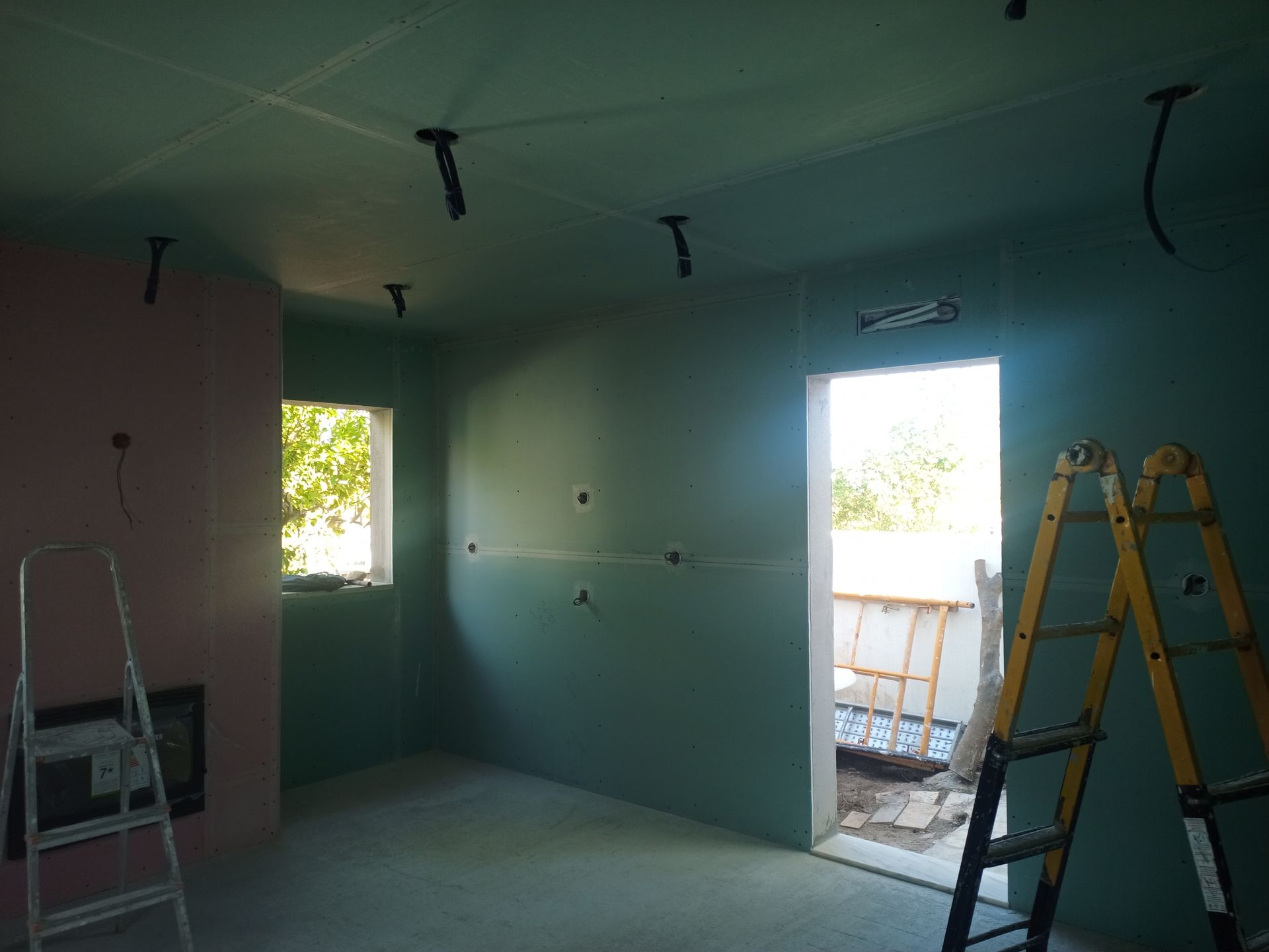 Cômodo interno em construção, com drywall verde, fios elétricos expostos, escadas e uma porta aberta.