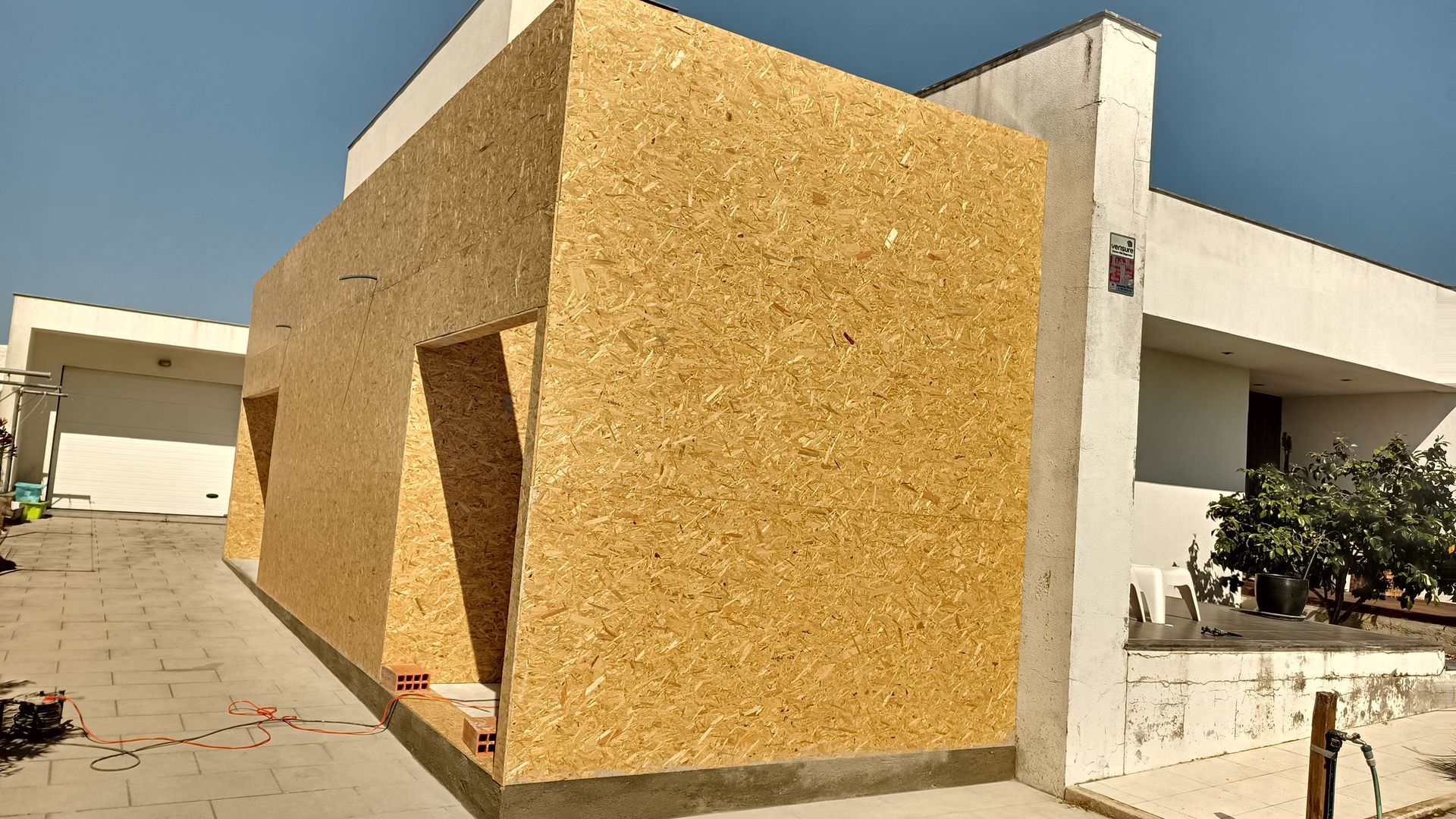 Vista exterior de um edifício em construção, com revestimento em aglomerado de madeira e portas abertas contra um céu azul brilhante.