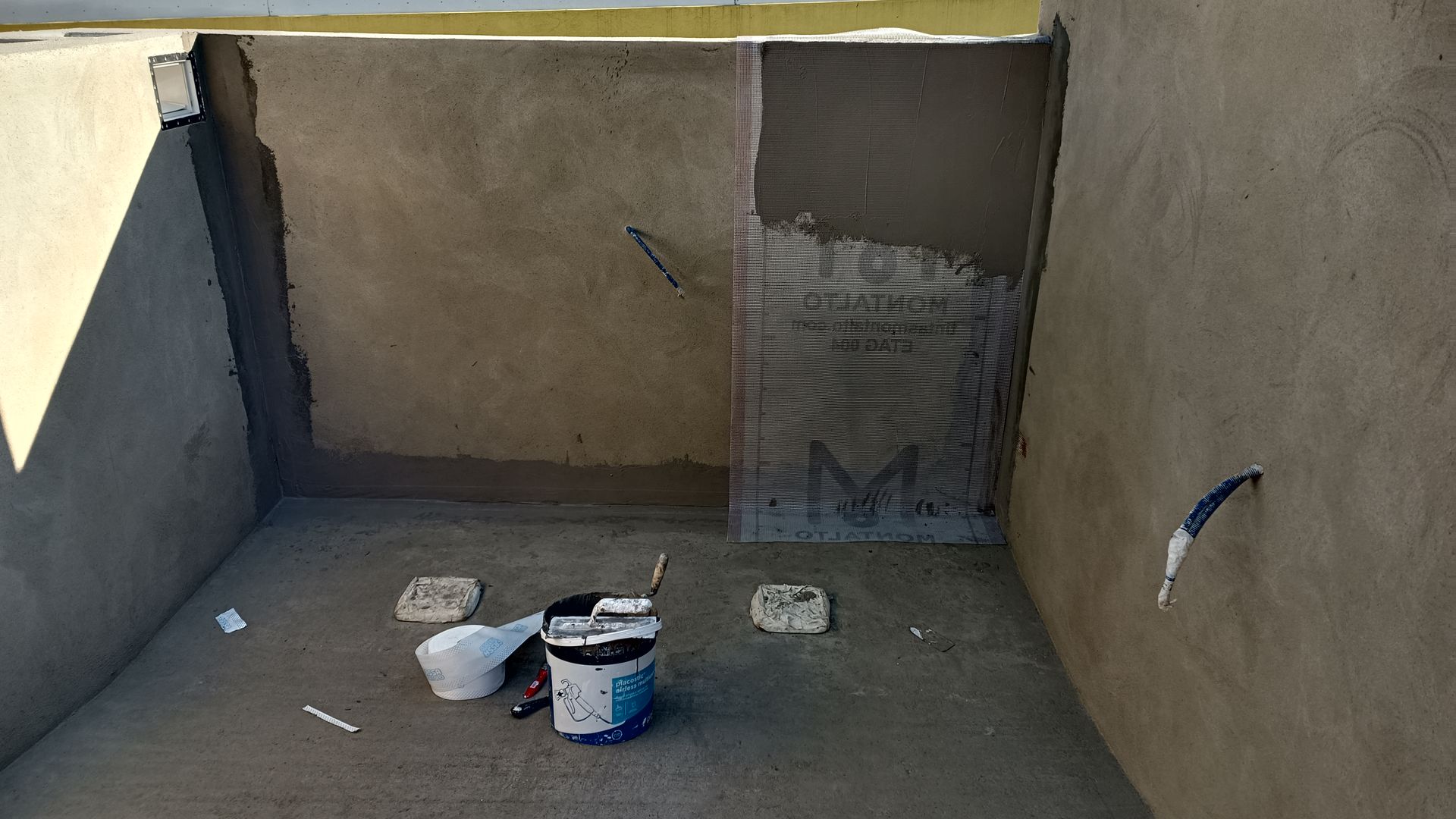 Interior de um cômodo parcialmente rebocado; piso e paredes de concreto cinza. Balde de tinta e materiais de pintura visíveis.