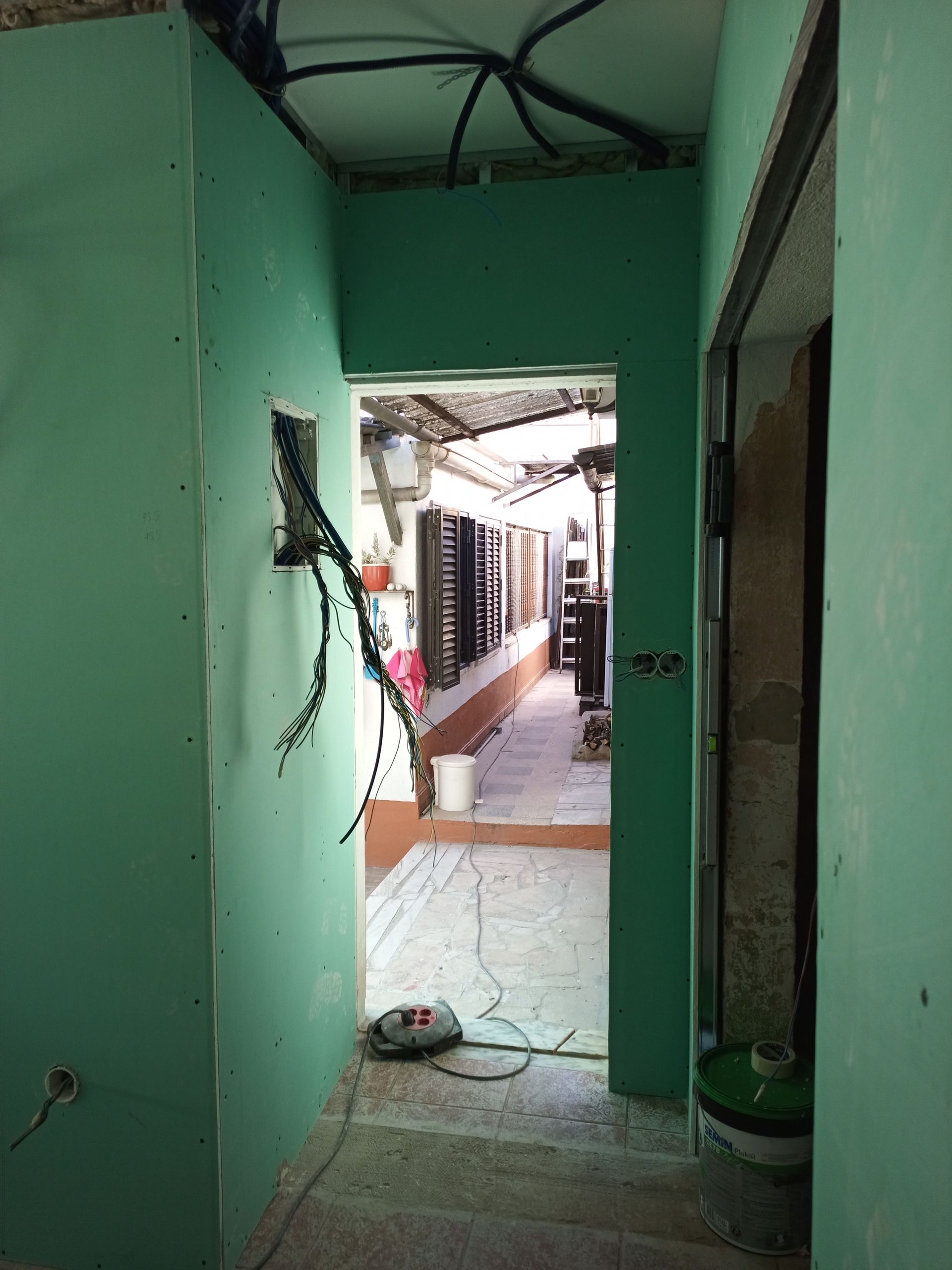 Corredor em construção, paredes verdes, porta para área externa com luz solar, fiação exposta.