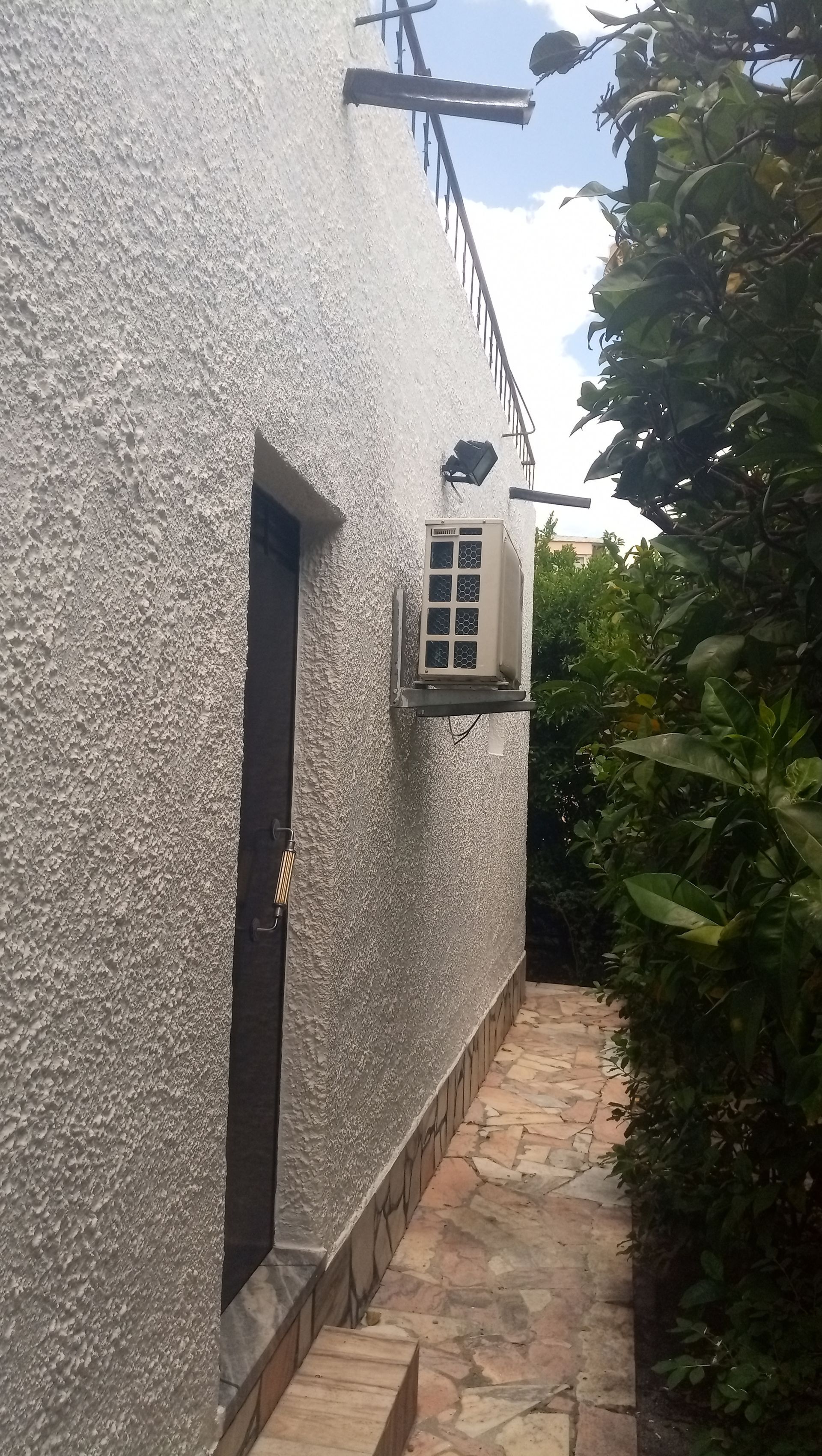 Parede externa com porta, ar condicionado e caminho de acesso. Árvores e antena na lateral.