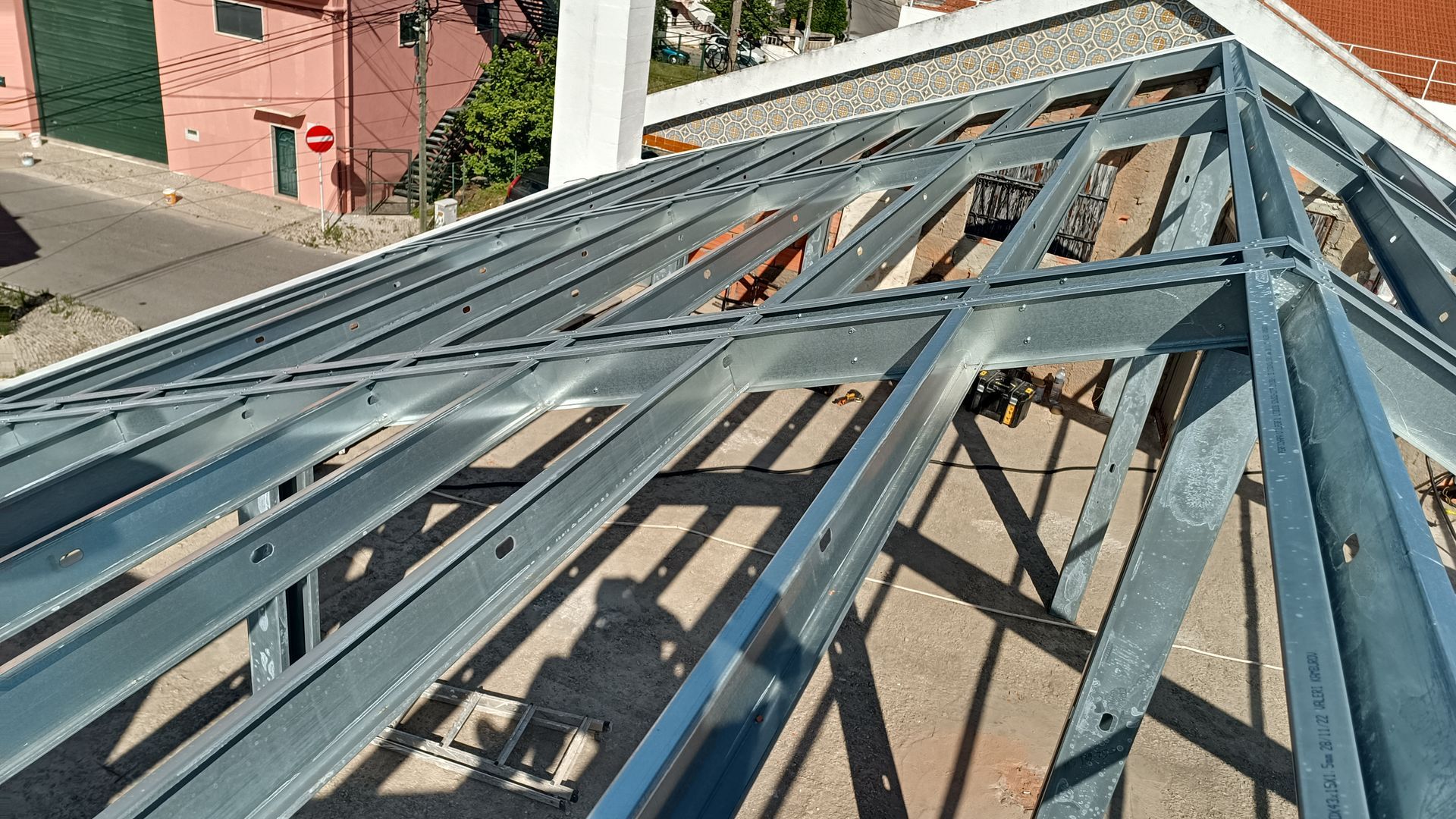 Estrutura metálica do telhado de um edifício. Vista de cima, com vigas expostas em padrão quadriculado. Área externa.