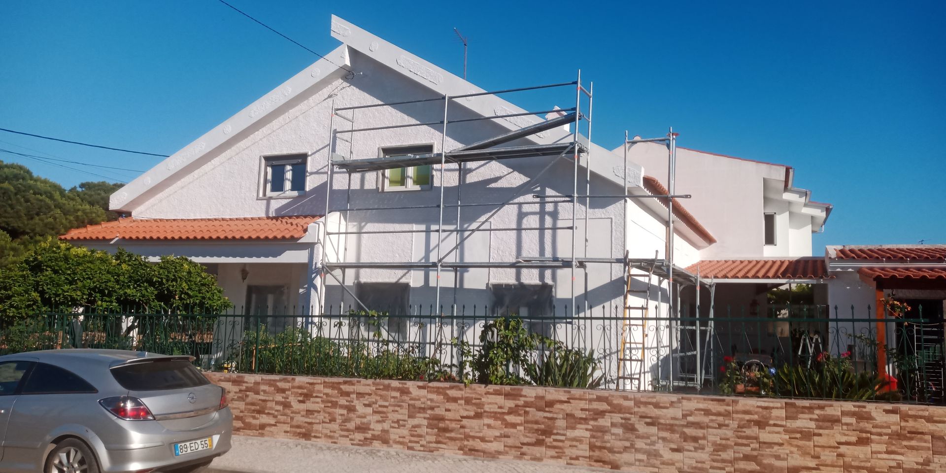Uma casa branca de dois andares em reforma, com andaimes montados, um carro estacionado nas proximidades e um céu azul claro.