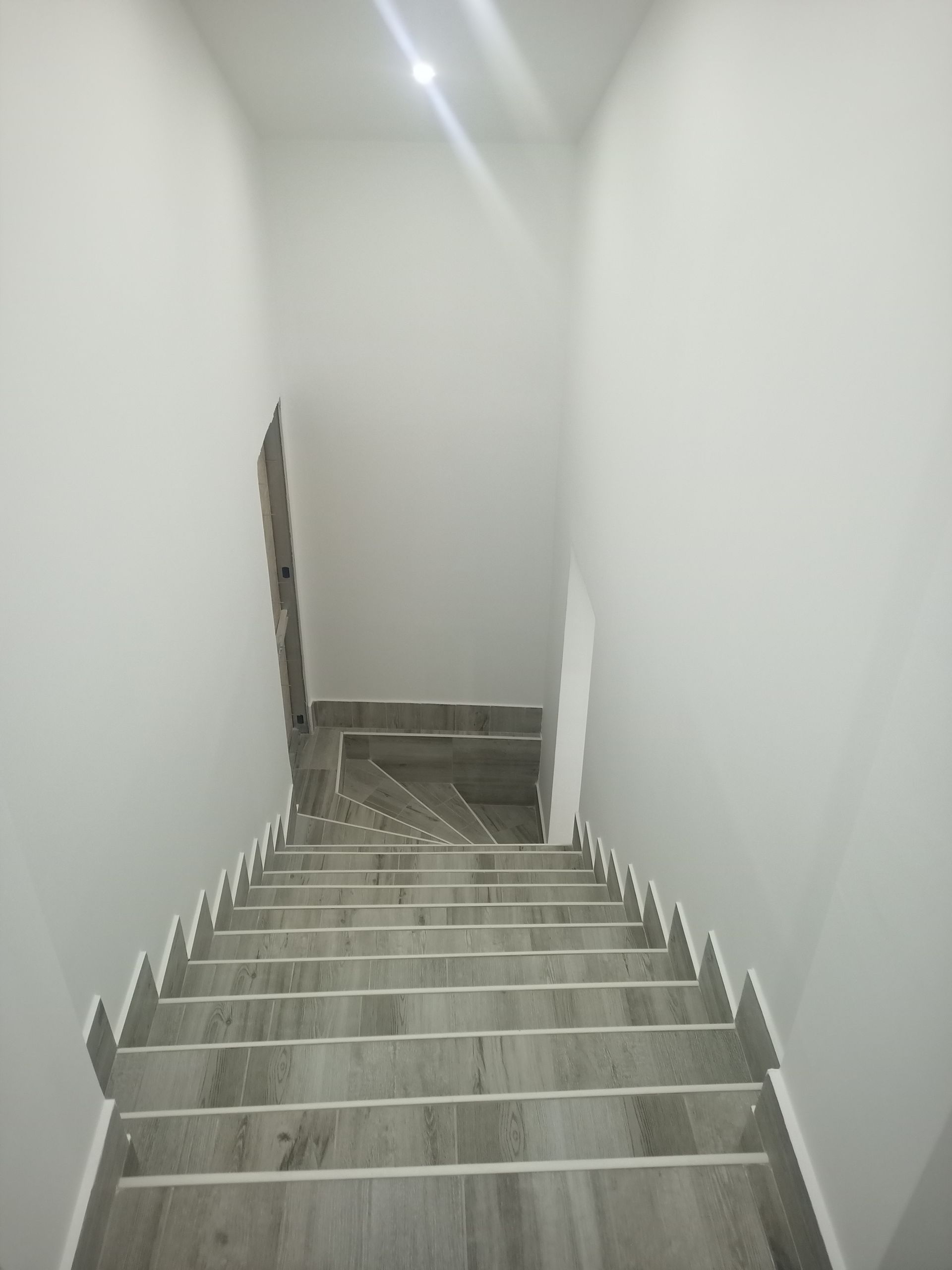 Escadaria descendente com degraus de azulejo cinza e paredes brancas. A luz incide do alto.