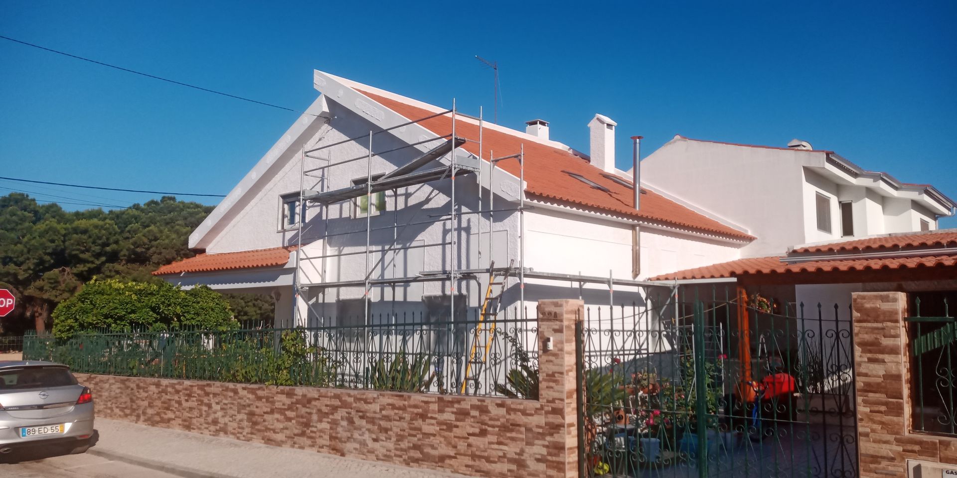 Casa branca com telhado de telha vermelha, andaimes e muro de tijolos em um dia ensolarado.